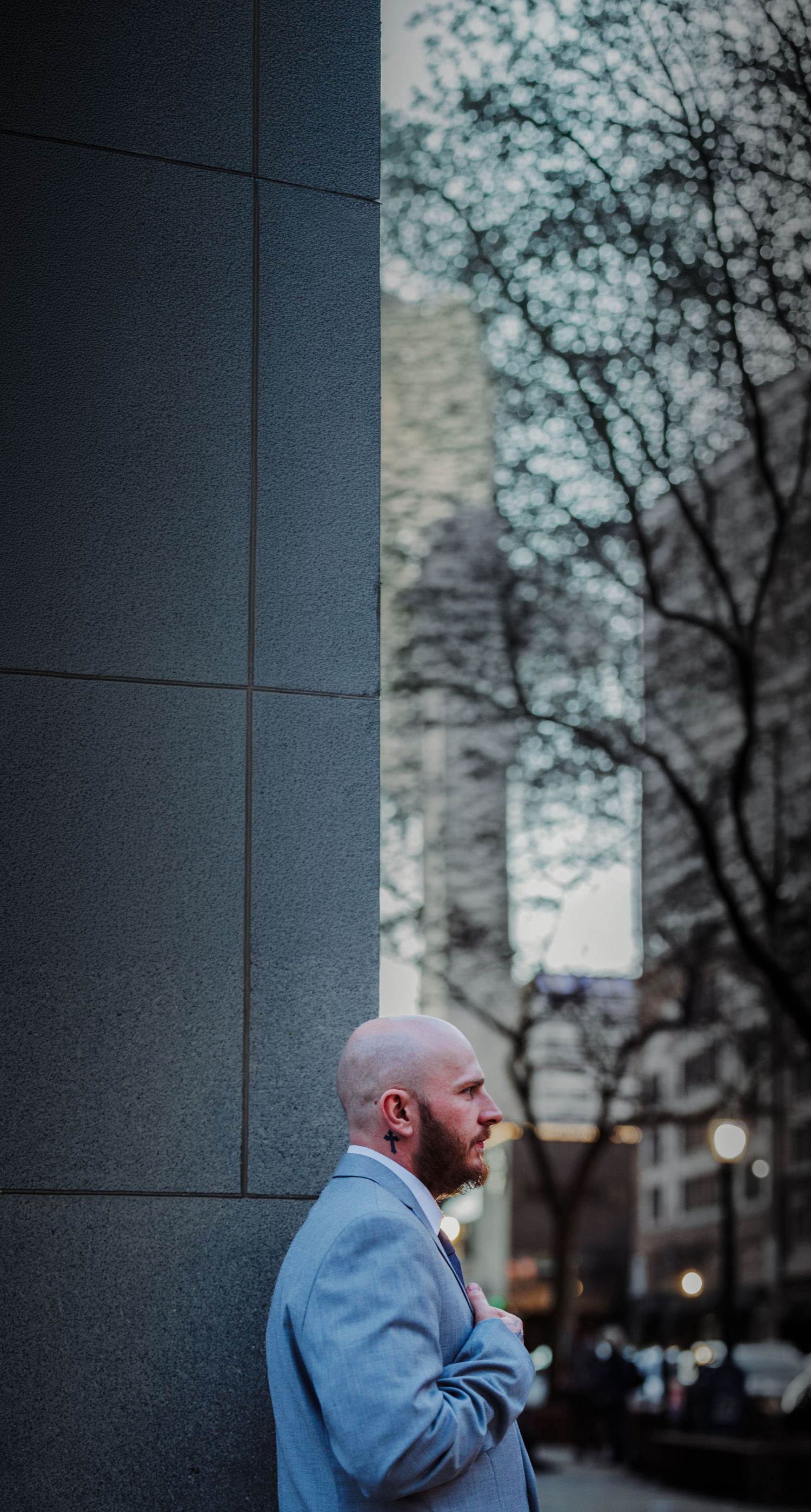 Bald bearded man in light blue suit leaning against stone building wall in urban setting with bare trees.