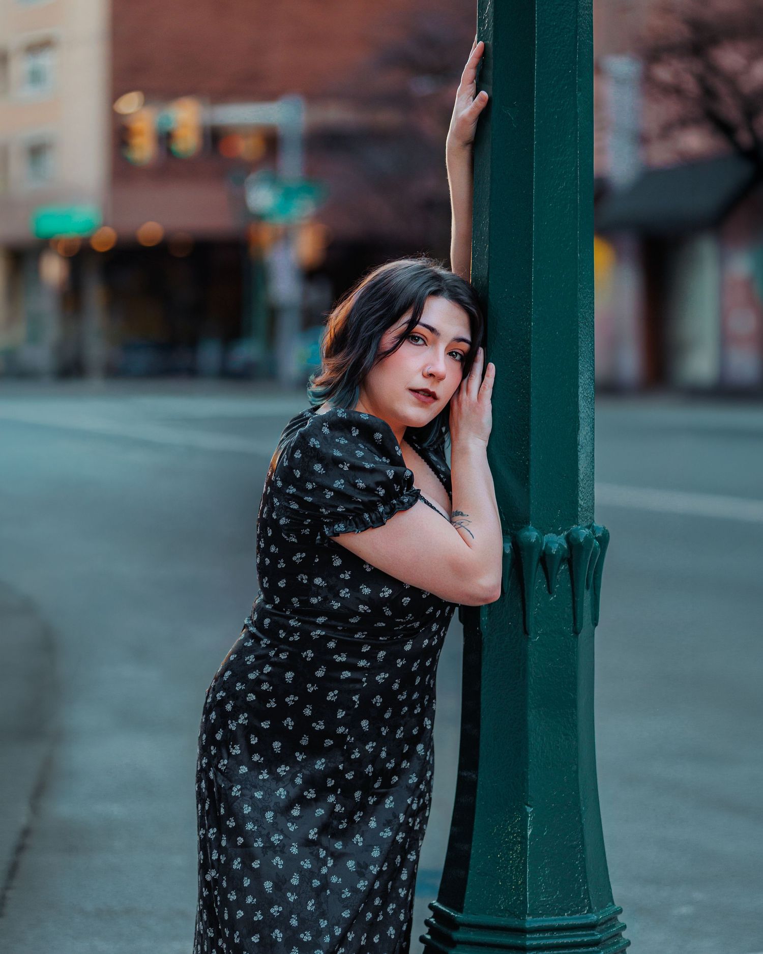 A young woman in a black floral dress leans against a green street lamp on an urban sidewalk at dusk.