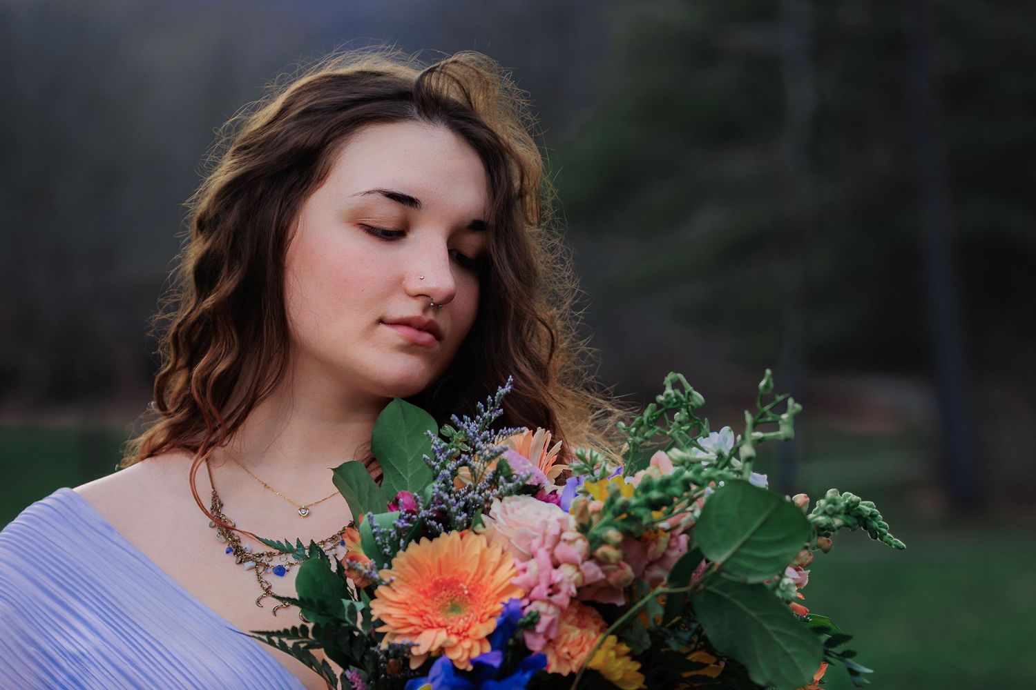Young woman with curly hair holding a colorful bouquet of flowers outdoors in a serene forest setting.