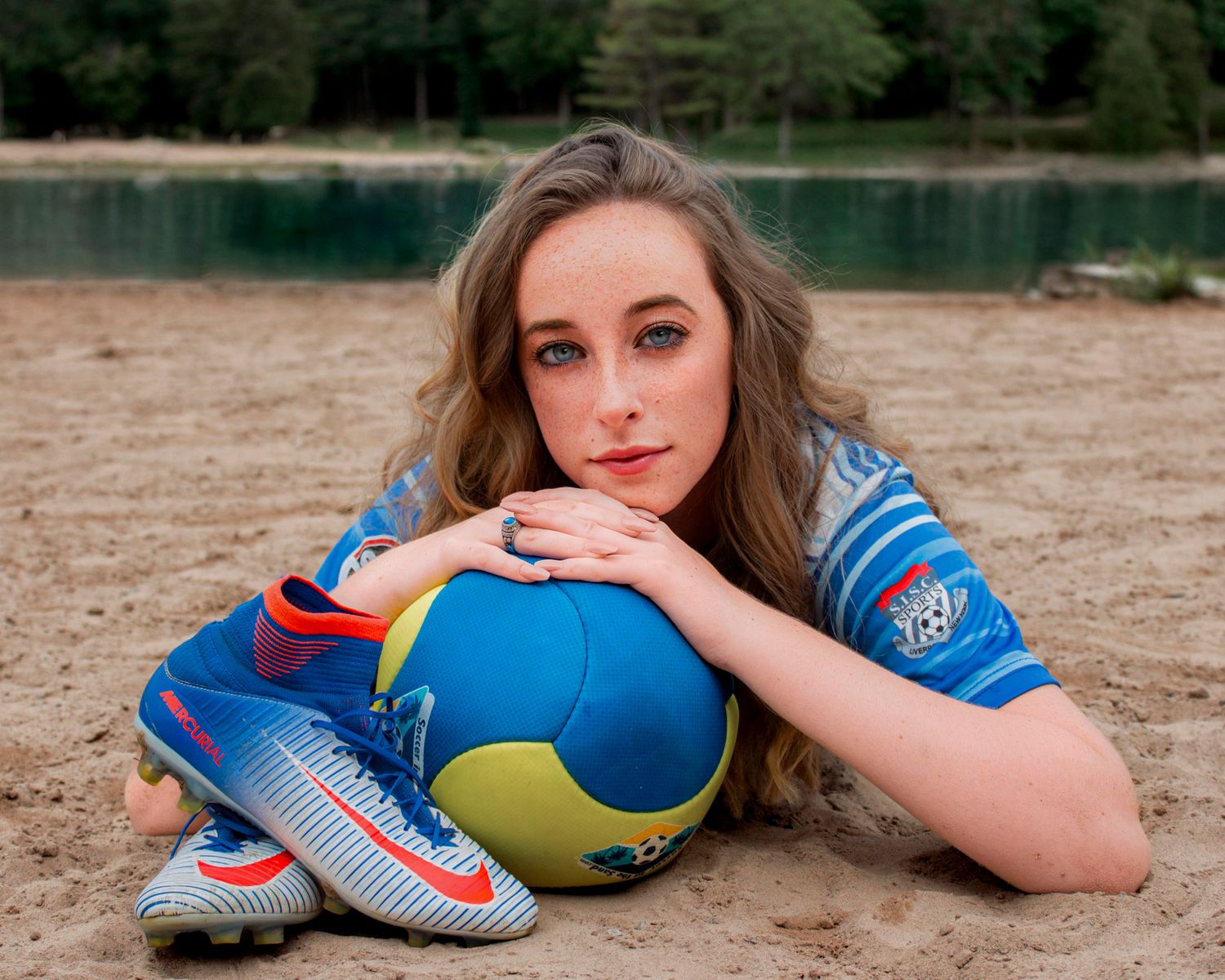 Young female soccer player lying on sandy beach with colorful ball and Nike cleats, wearing blue jersey.