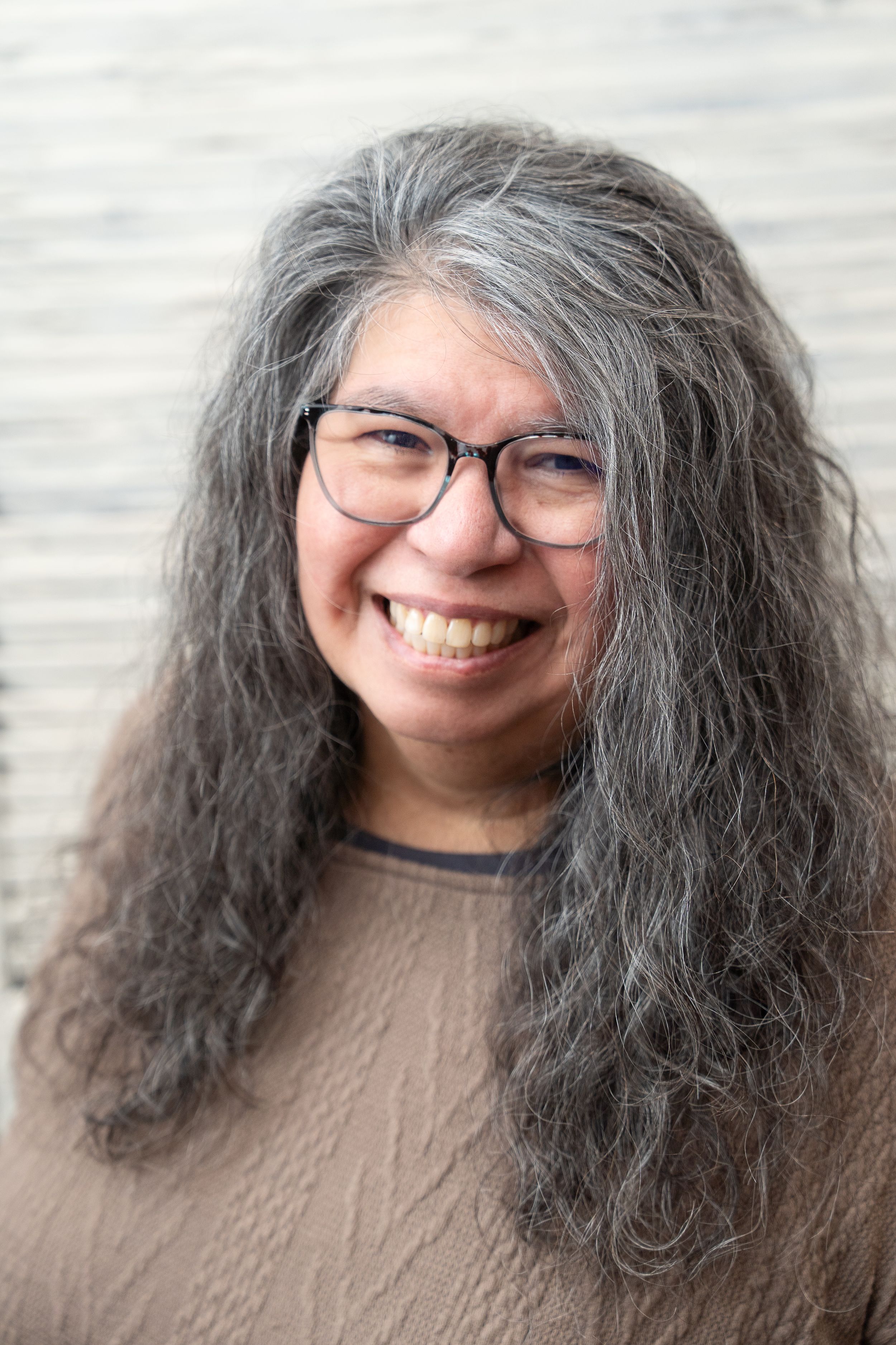 A portrait showing a genuine warm smile, glasses, and long flowing grey hair against a light background.