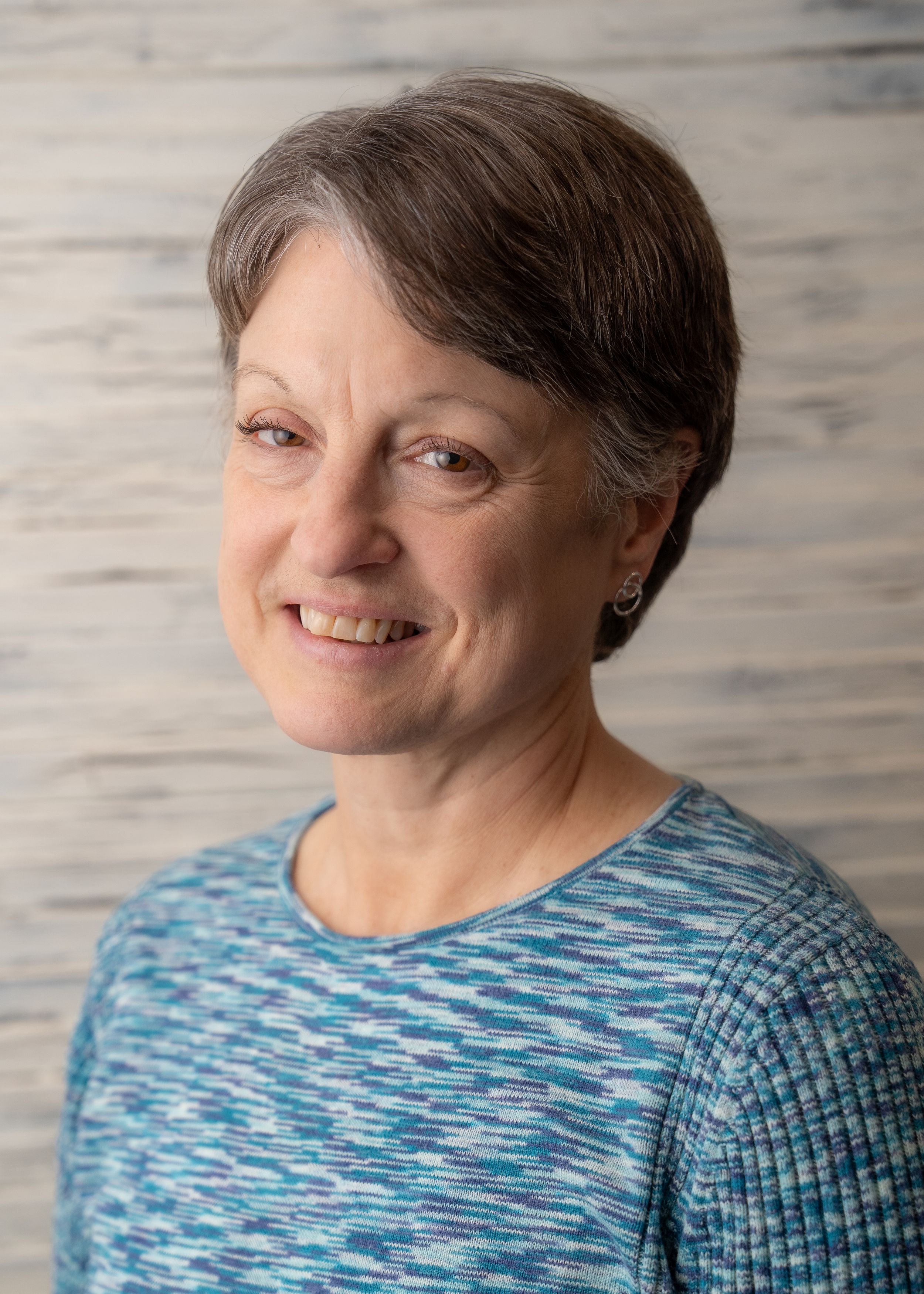 A person wearing a blue marbled sweater smiles warmly against a textured grey background.
