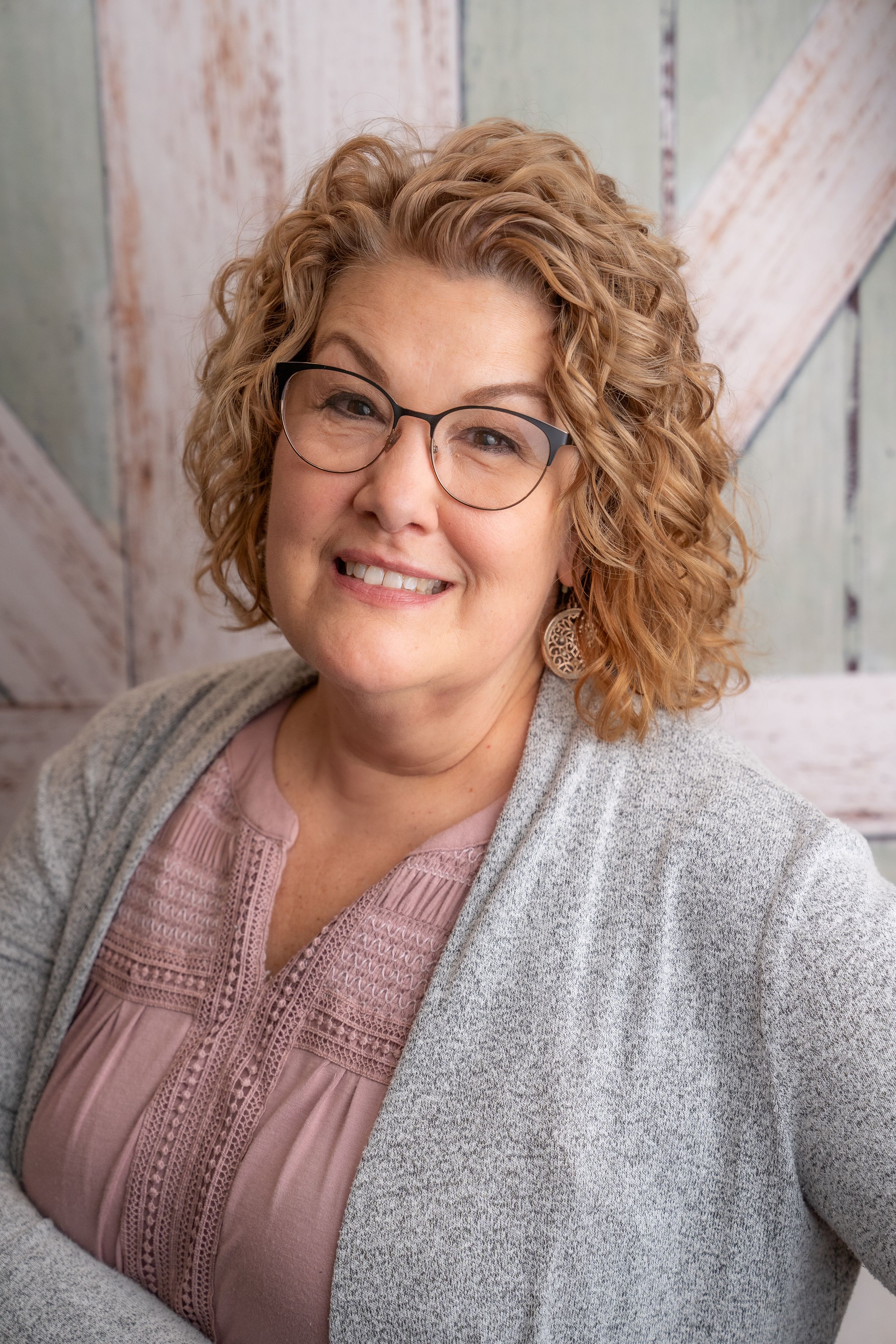 Professional headshot of a person with curly blonde hair wearing glasses, a pink blouse and grey cardigan against a wooden backdrop.