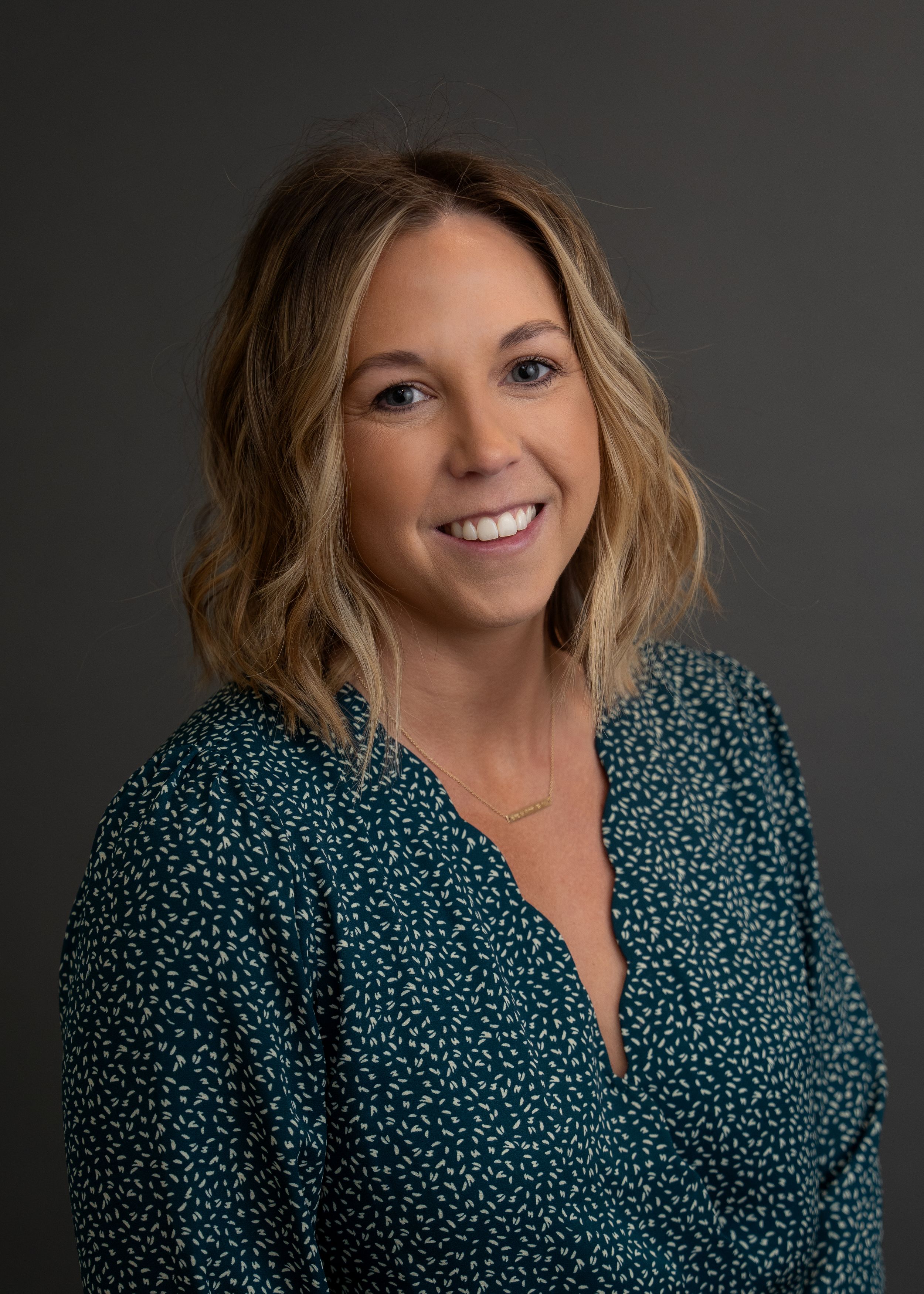 Professional headshot of someone in a dark teal patterned wrap blouse against a dark gray background.
