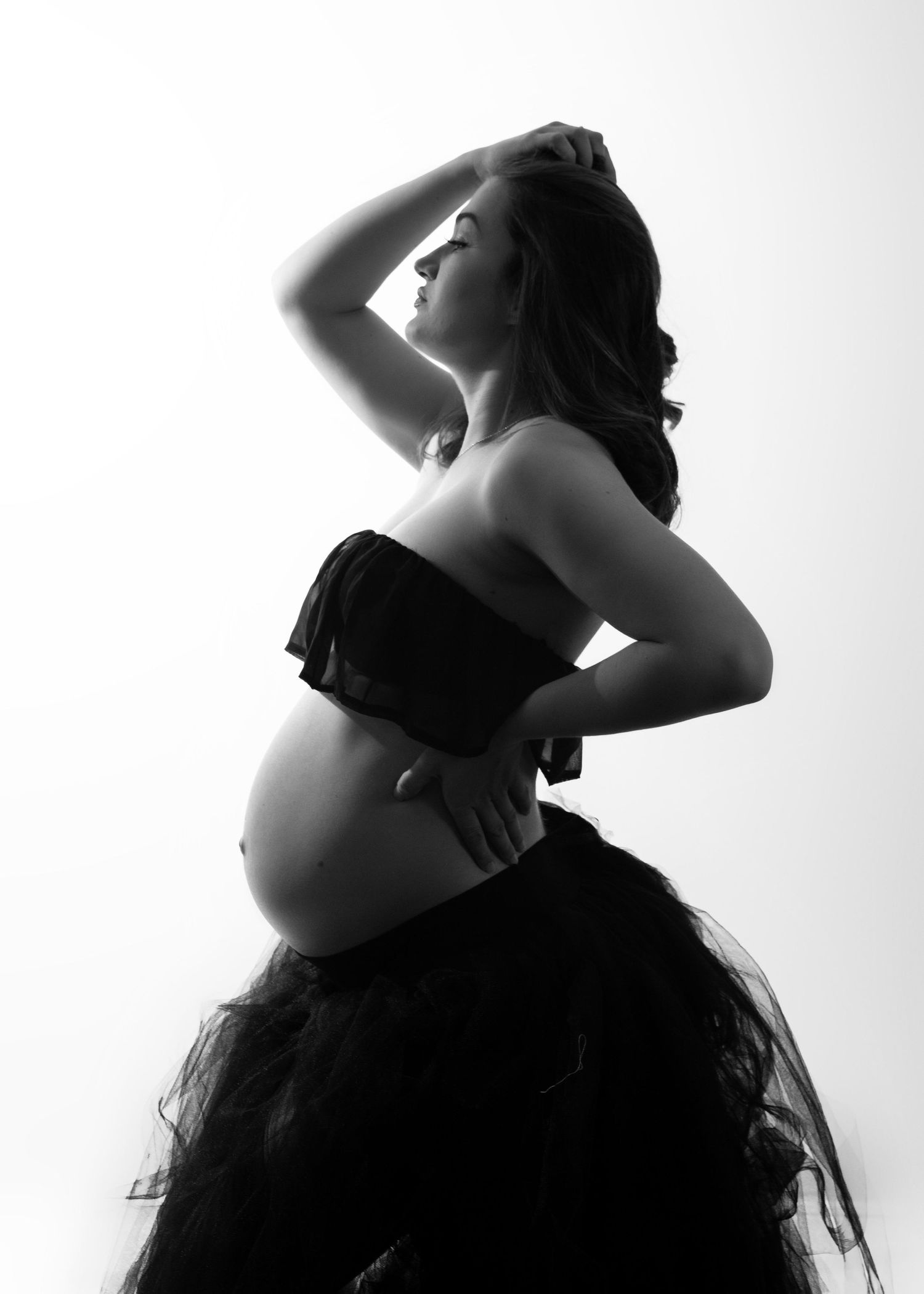 Artistic black and white maternity photo showing silhouette of pregnant woman in tulle skirt against light background.