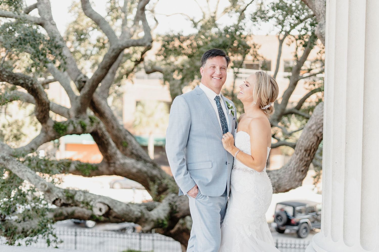 A couple in wedding attire pose together under sprawling oak trees at a sunny outdoor venue.