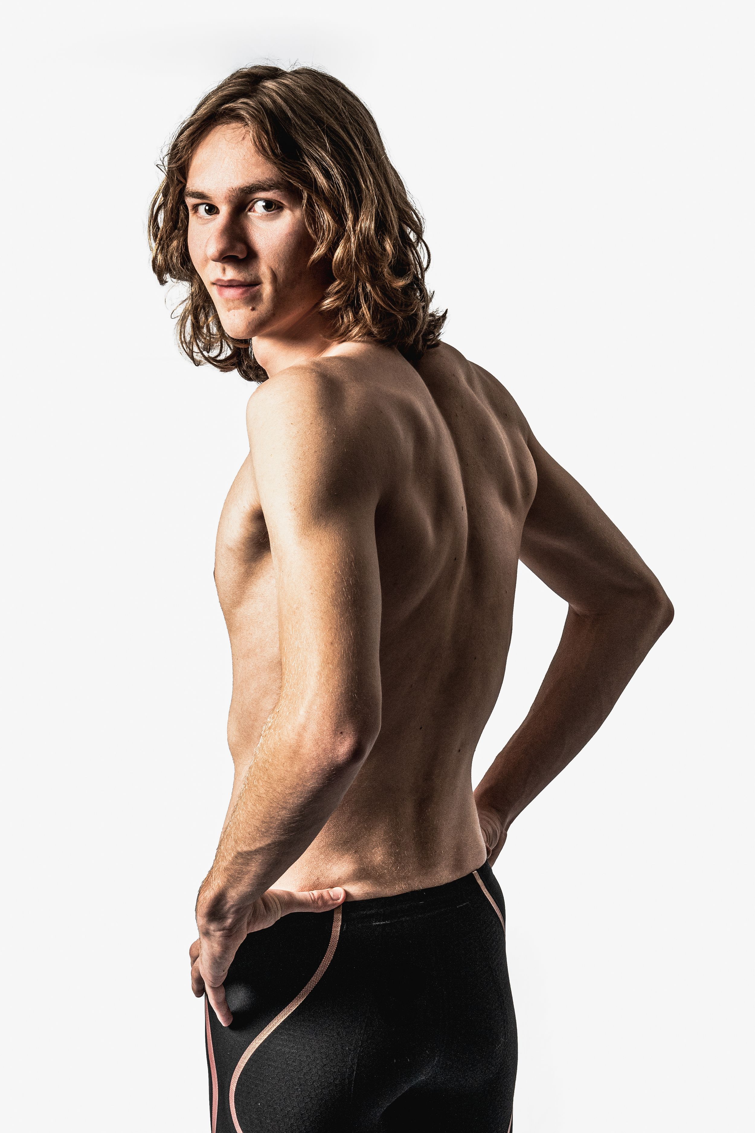 Athletic person with long hair poses shirtless against white background showing muscular back and shoulders.