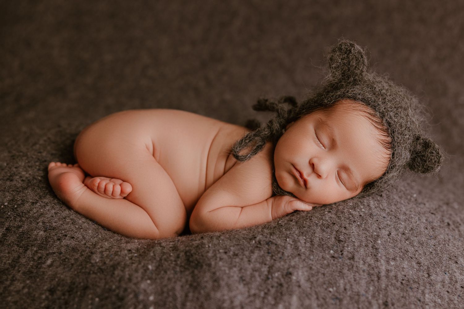 A peaceful sleeping newborn wearing a cozy knit bear bonnet on textured brown fabric.