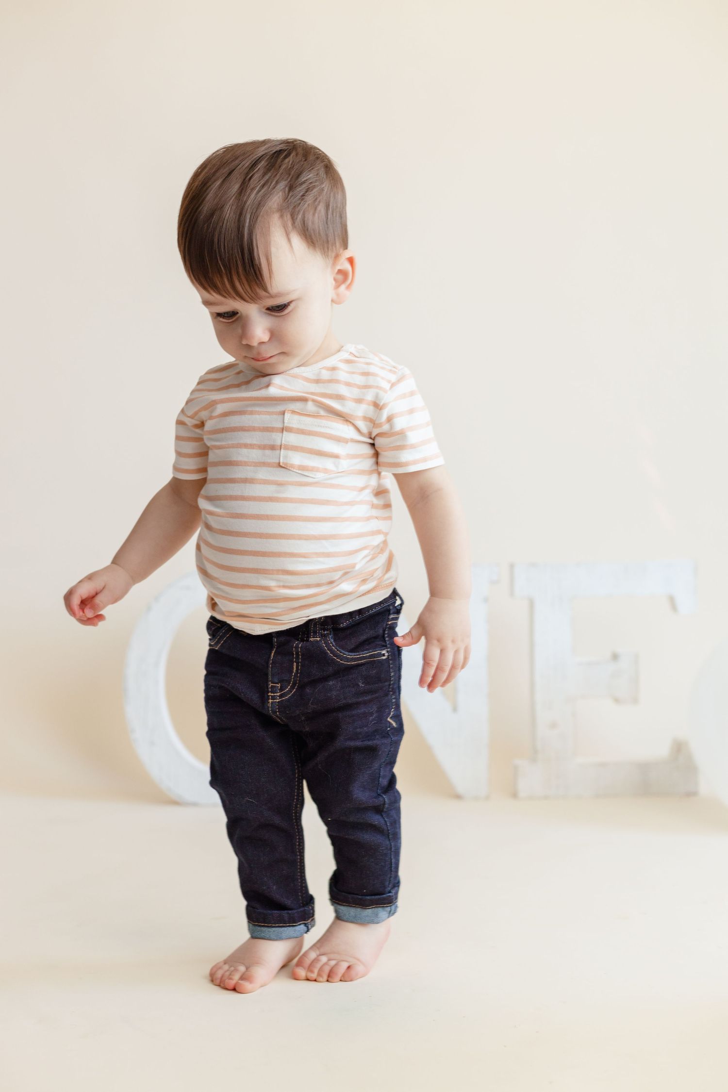 A toddler in a striped shirt and jeans stands barefoot against a light background.