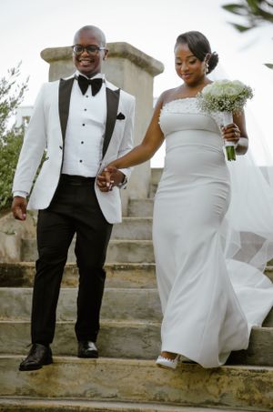 Elegant couple on stone steps at their wedding ceremony, with bride in white dress holding bouquet and groom in white tuxedo jacket.