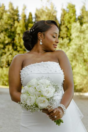 Elegant bride in white strapless gown holds a stunning bouquet of white roses and baby's breath against sunlit greenery.