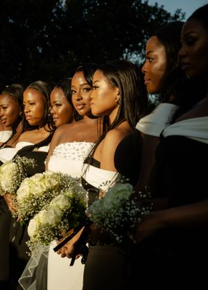 Wedding party wearing black bridesmaid dresses and a white bridal gown pose together holding baby's breath bouquets at sunset.