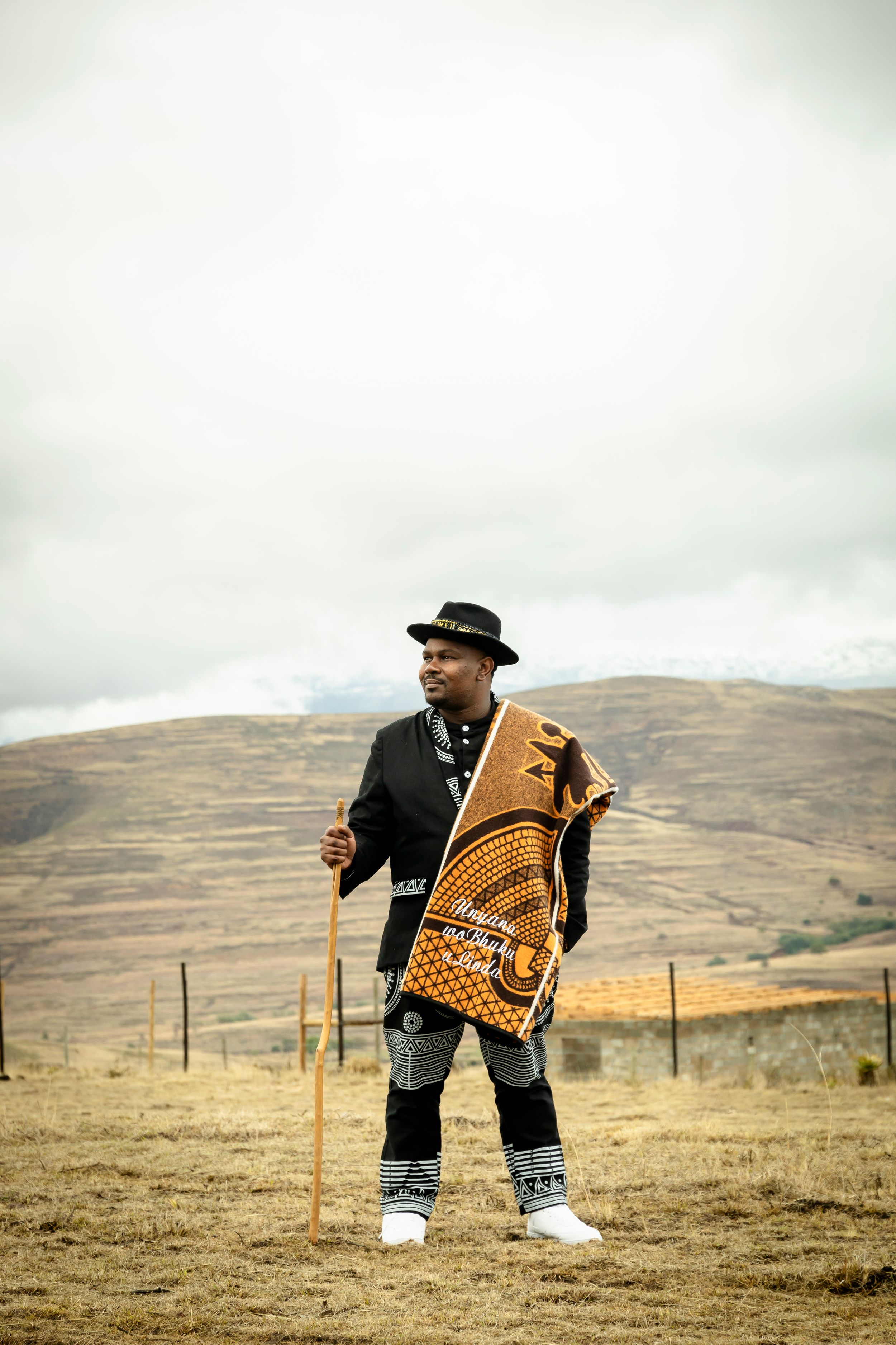 Person wearing hat and traditional blanket stands in mountainous terrain with walking stick on overcast day.