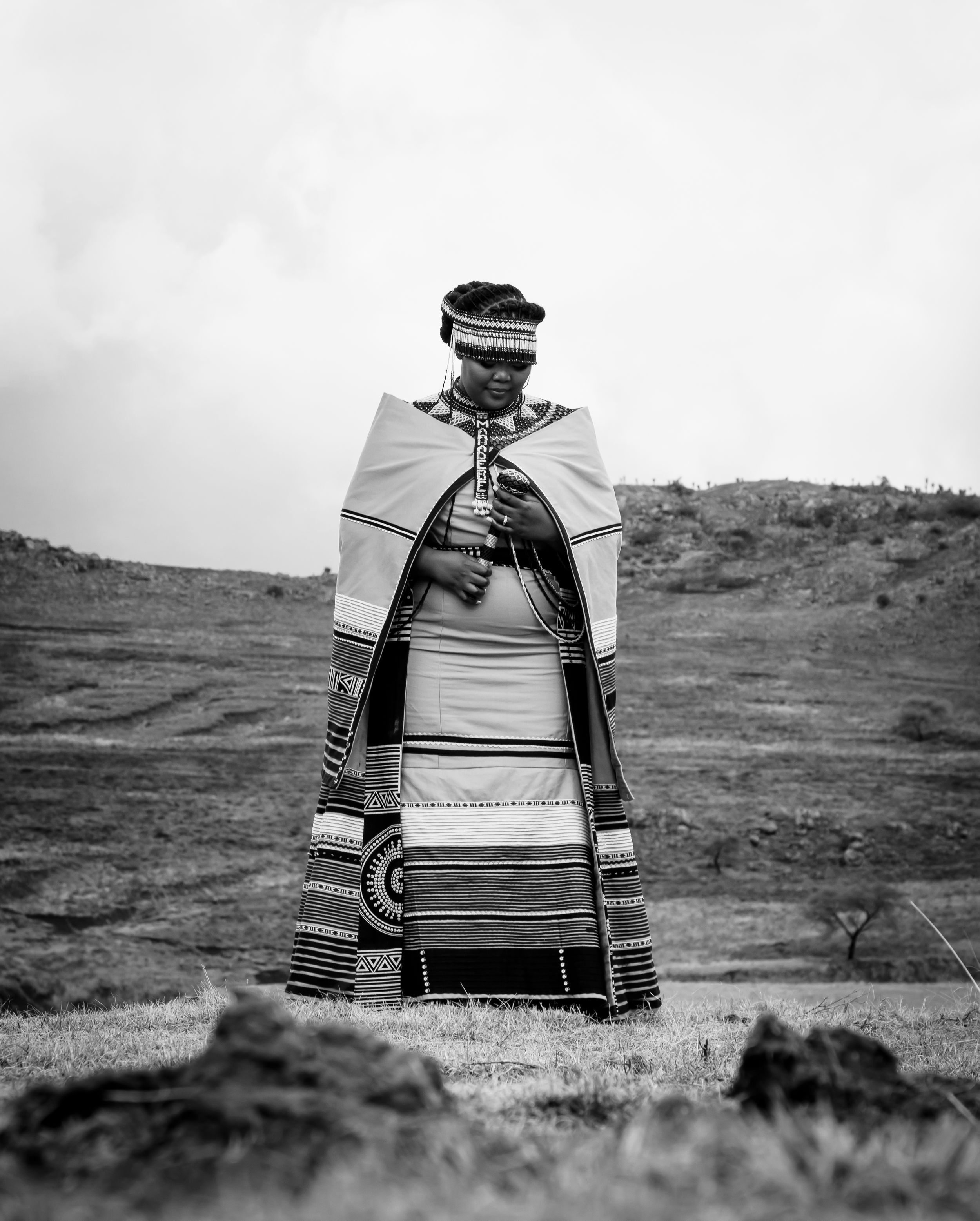 Black and white photograph of someone wearing traditional Xhosa attire with blankets and decorative stripes standing in a field.