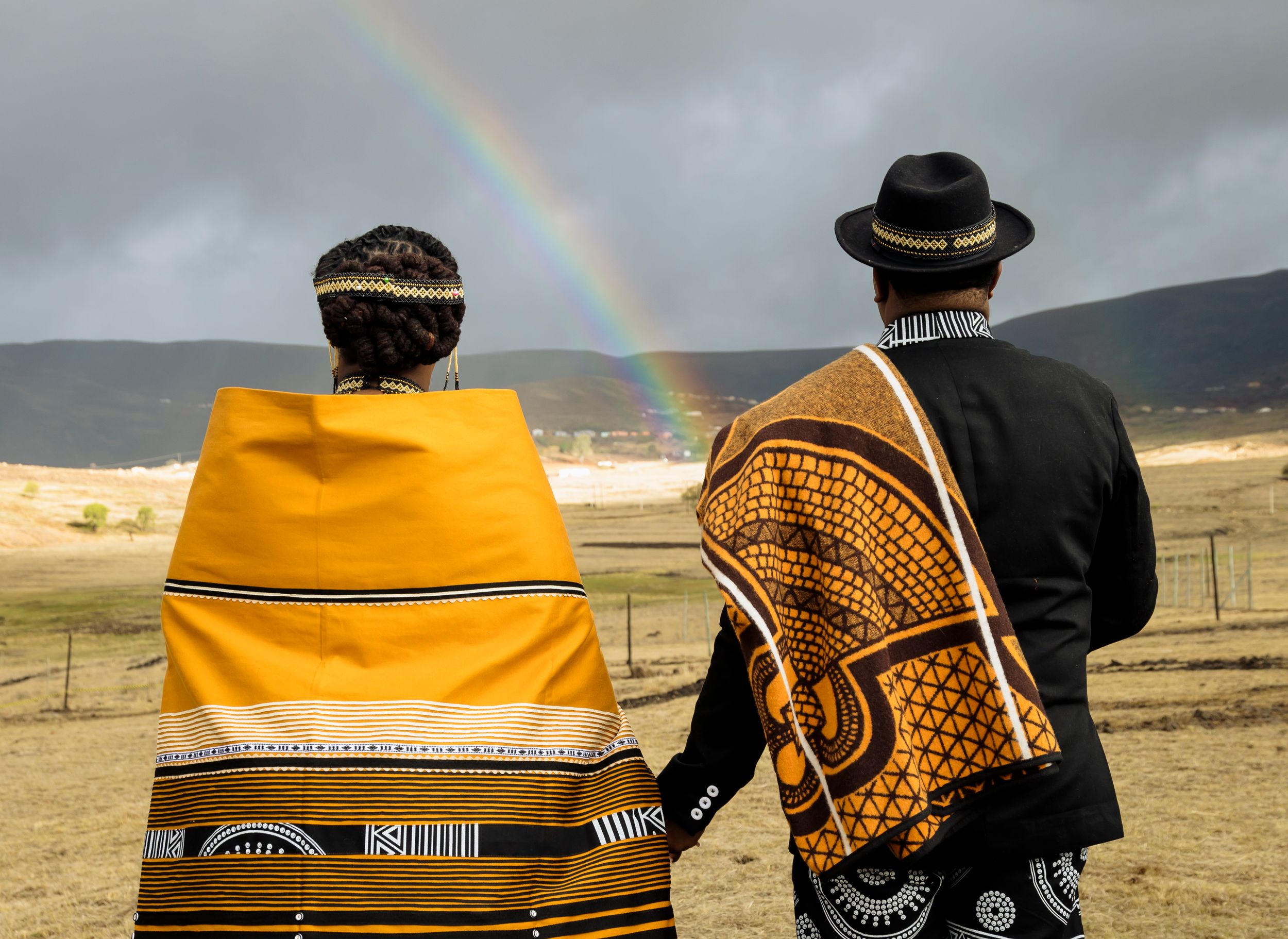 Two people in traditional clothing watch a rainbow over a desert landscape while sitting on a wooden fence.