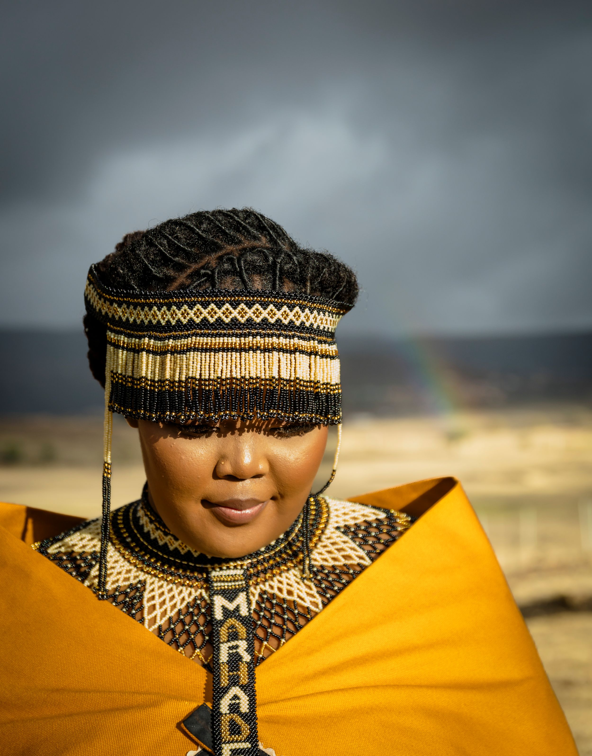 A striking portrait in traditional African attire with golden fabric and beaded headpiece against a stormy sky with rainbow.