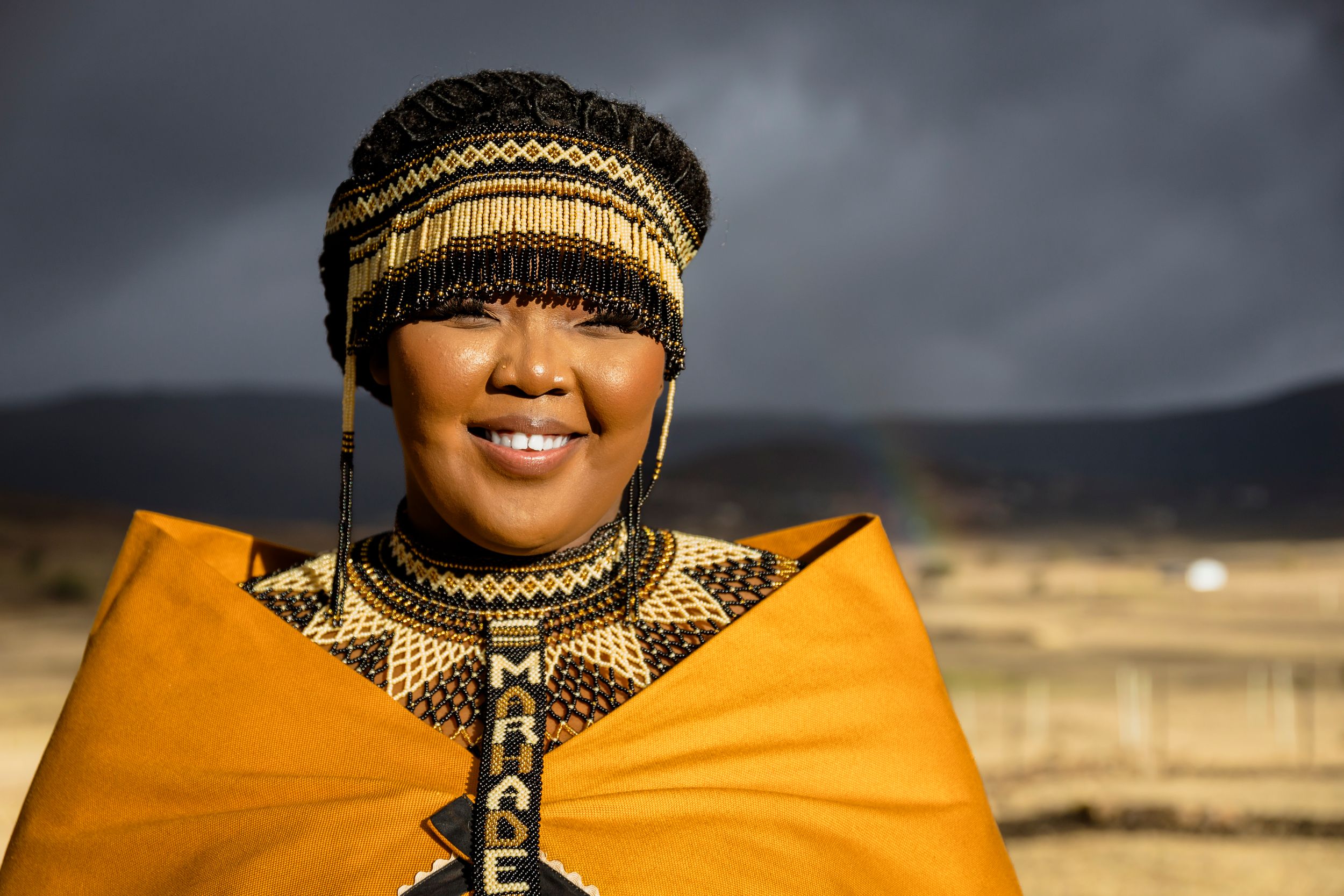 A person wearing traditional African attire with a golden cape and beaded headpiece smiles against a stormy sky background.