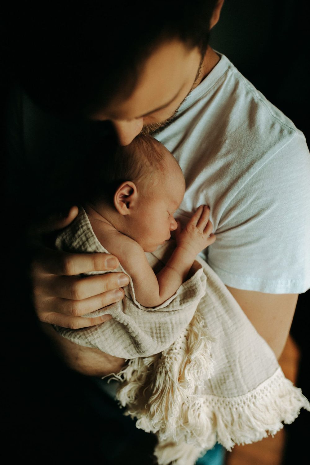 A tender moment captured in moody lighting as a sleeping baby in white lace clothing is held close against a white shirt.