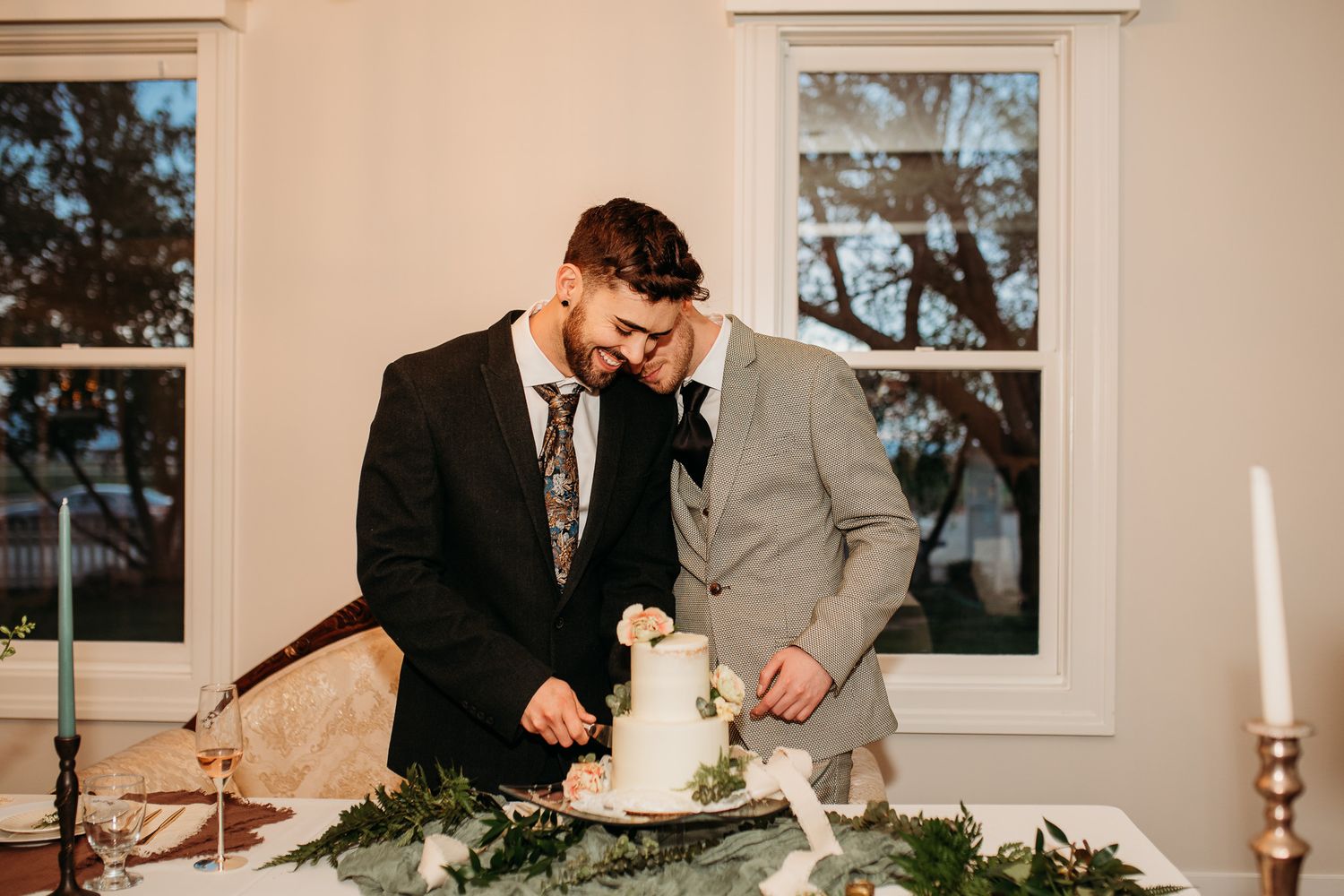 Two people in suits cutting their wedding cake together at a reception.