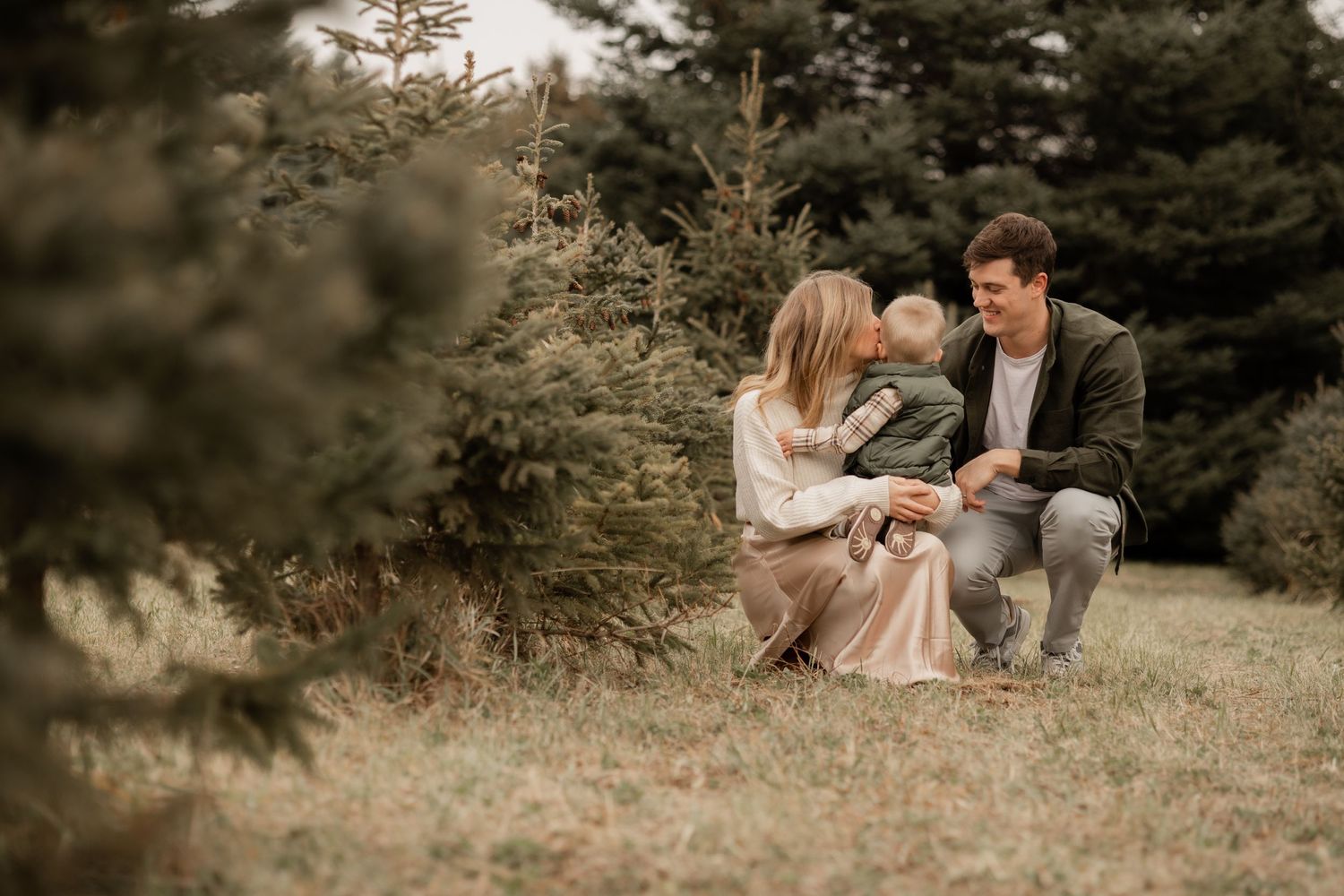 A couple sits together on grass surrounded by evergreen trees in a romantic outdoor setting.