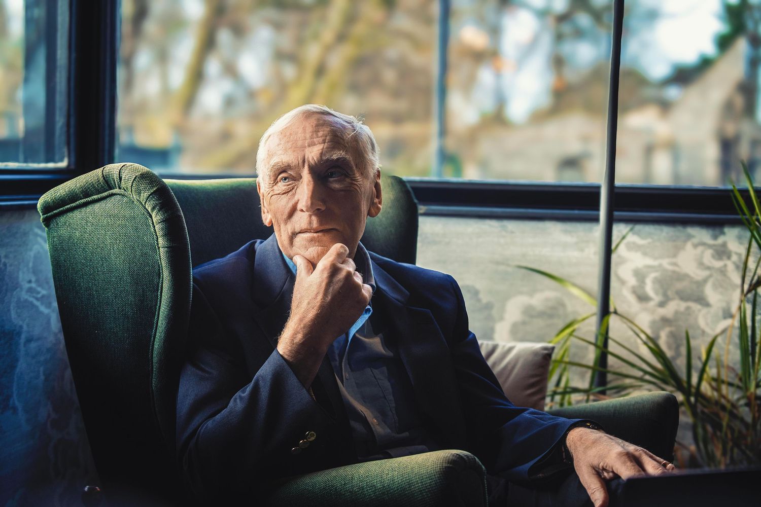 Contemplative senior executive sits in green armchair by window.