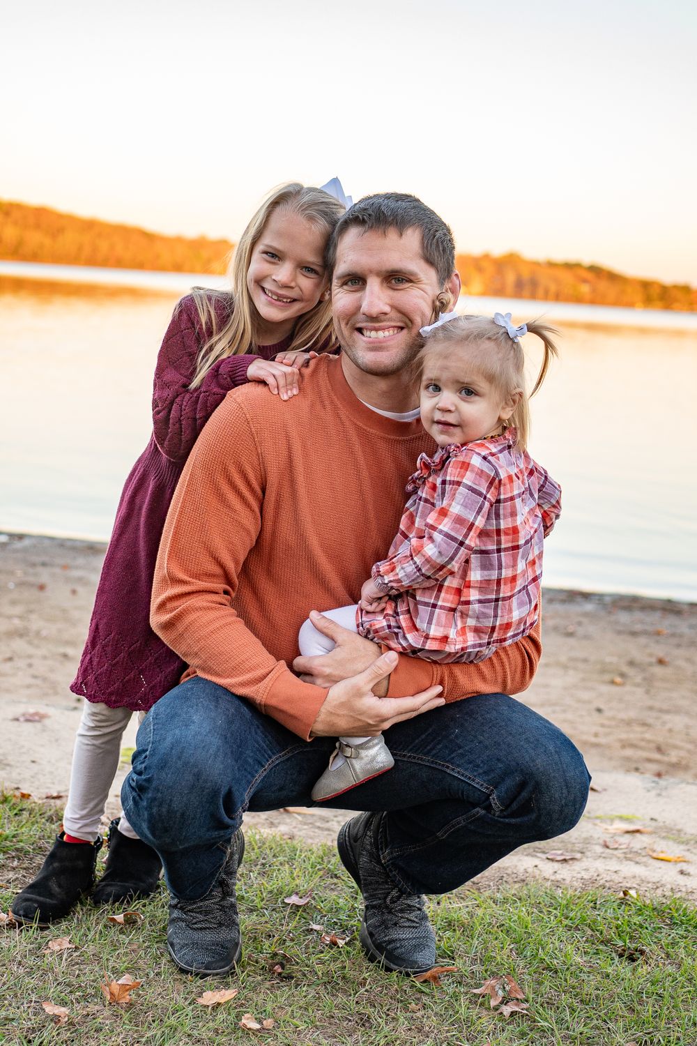Smiling father crouching by a lake at sunset, holding two young daughters in fall attire.