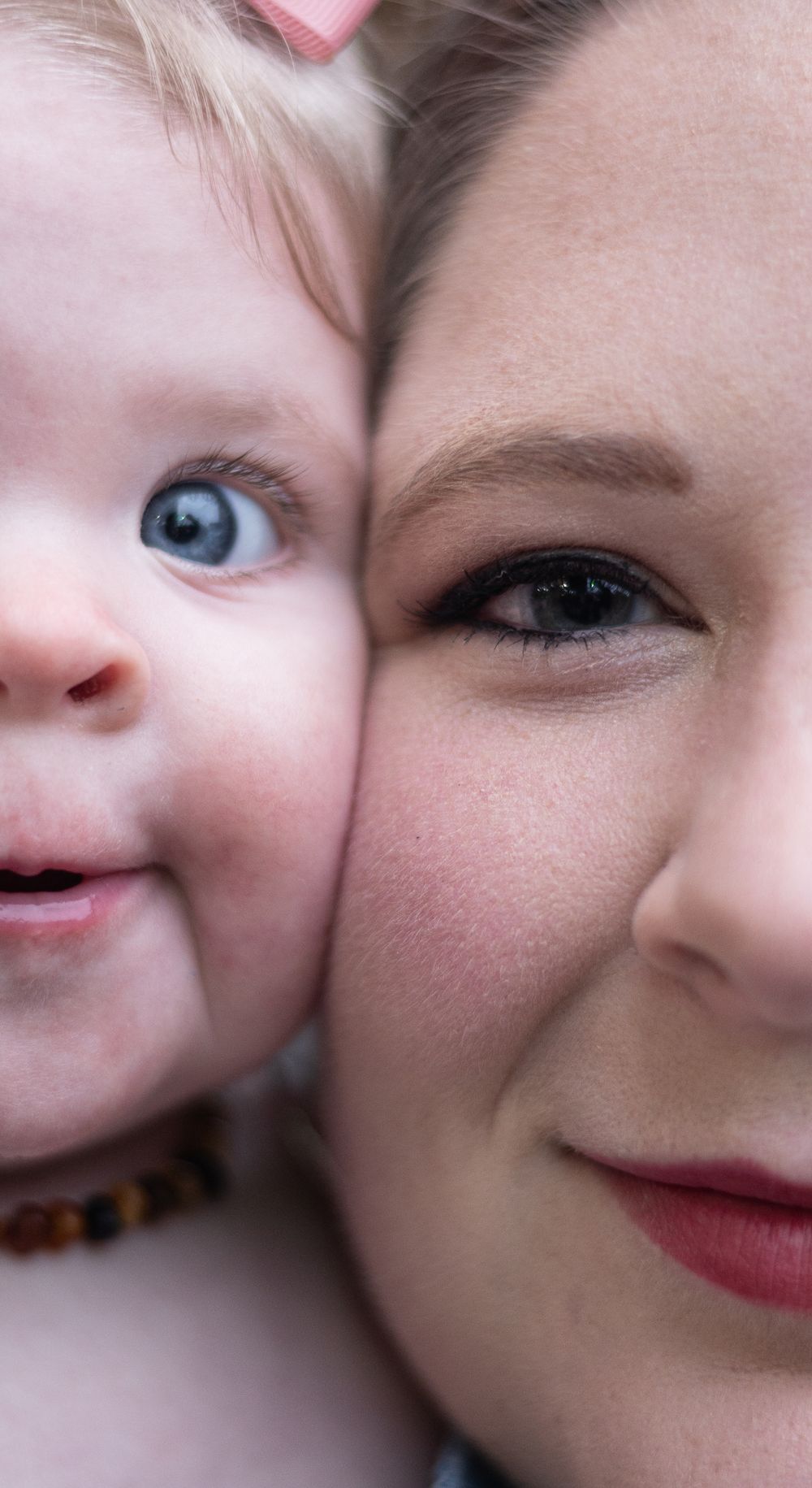 Close-up portrait of a smiling baby with blue eyes cheek-to-cheek with a young woman wearing red lipstick.