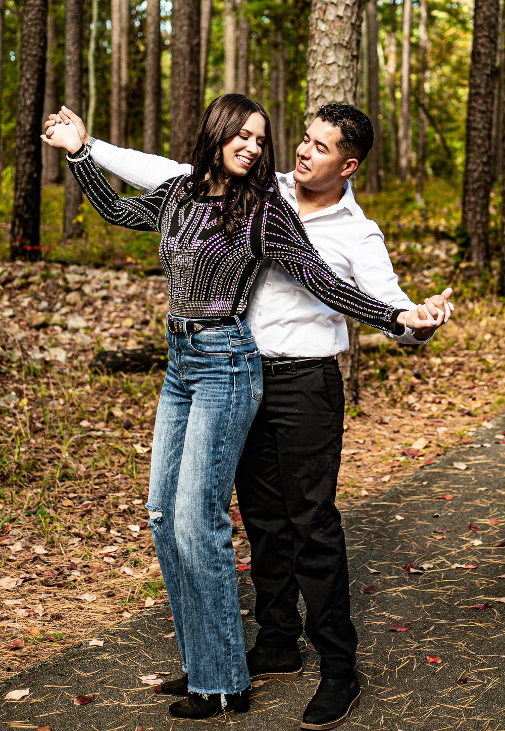 Couple dancing joyfully on a forest path, woman in sequined top and jeans, man in white shirt and black pants.