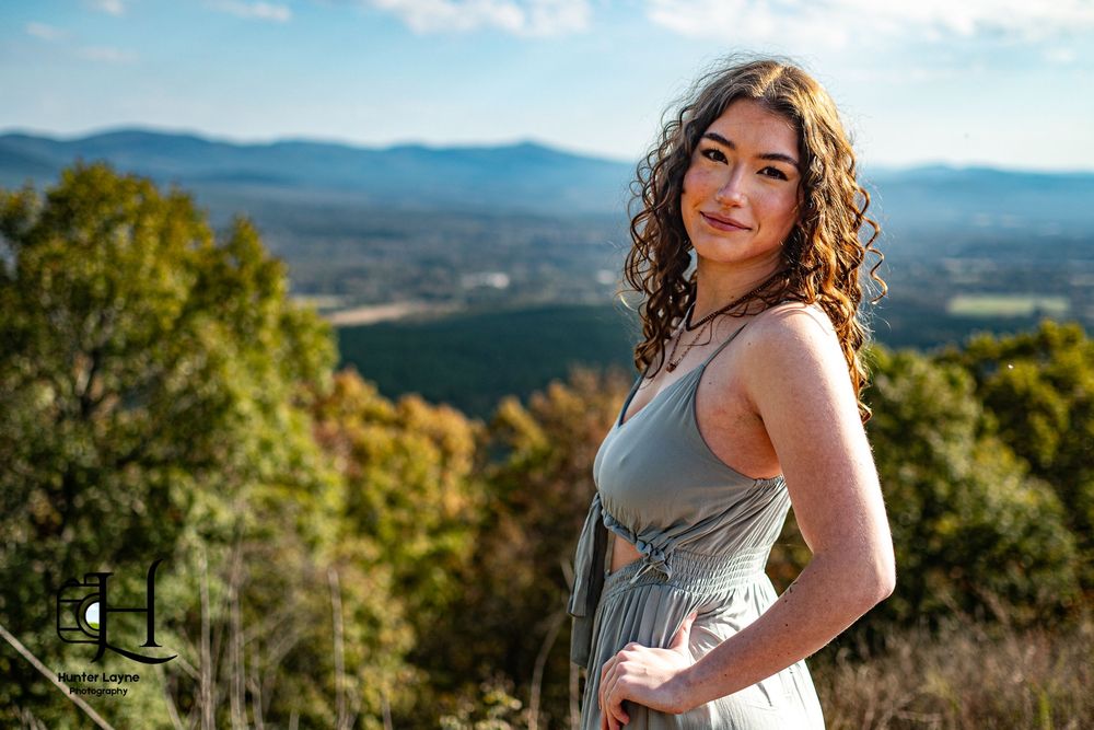 Young woman with curly hair in a gray dress posing on a hilltop with mountain views.