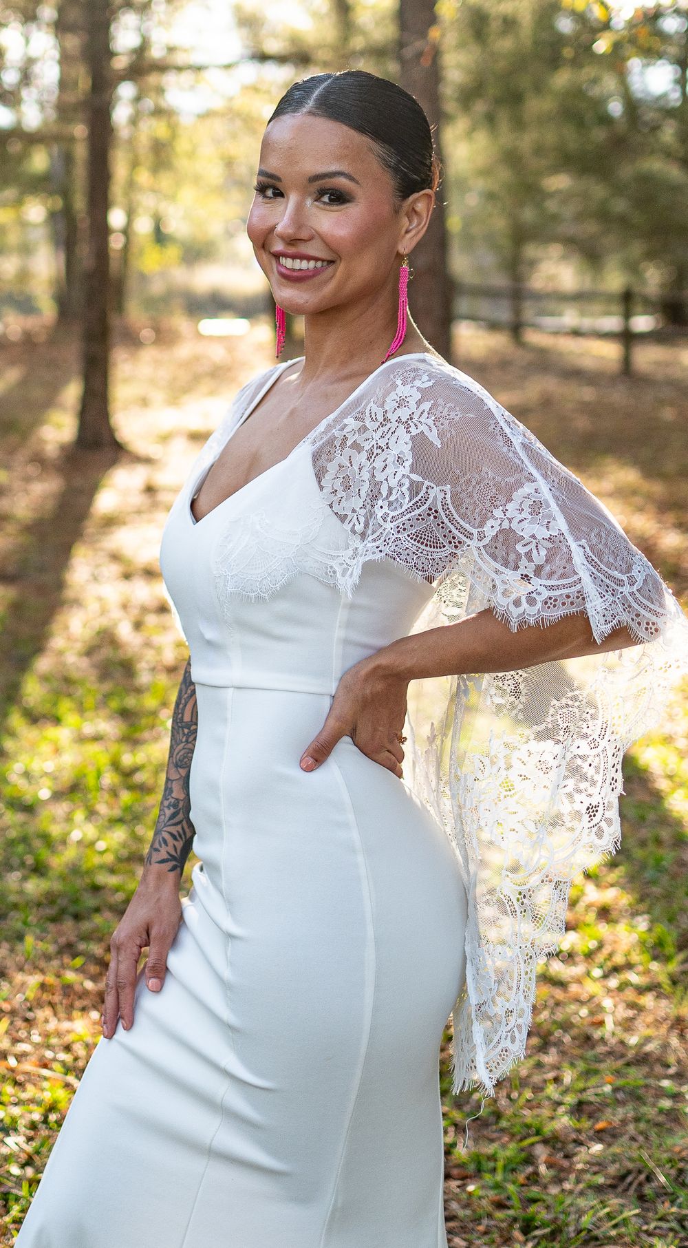 Smiling bride in a fitted white lace-sleeve wedding gown posing outdoors in sunlight.