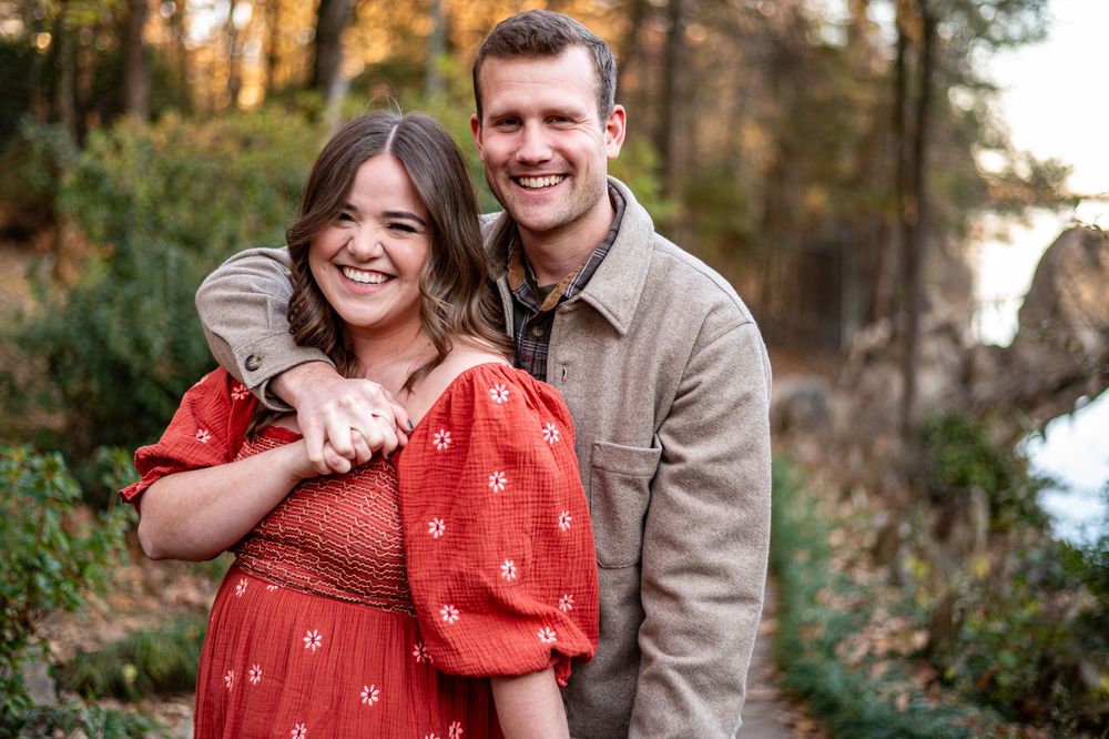 Happy couple embracing outdoors in autumn forest, woman in red floral dress, man in gray jacket, smiling.