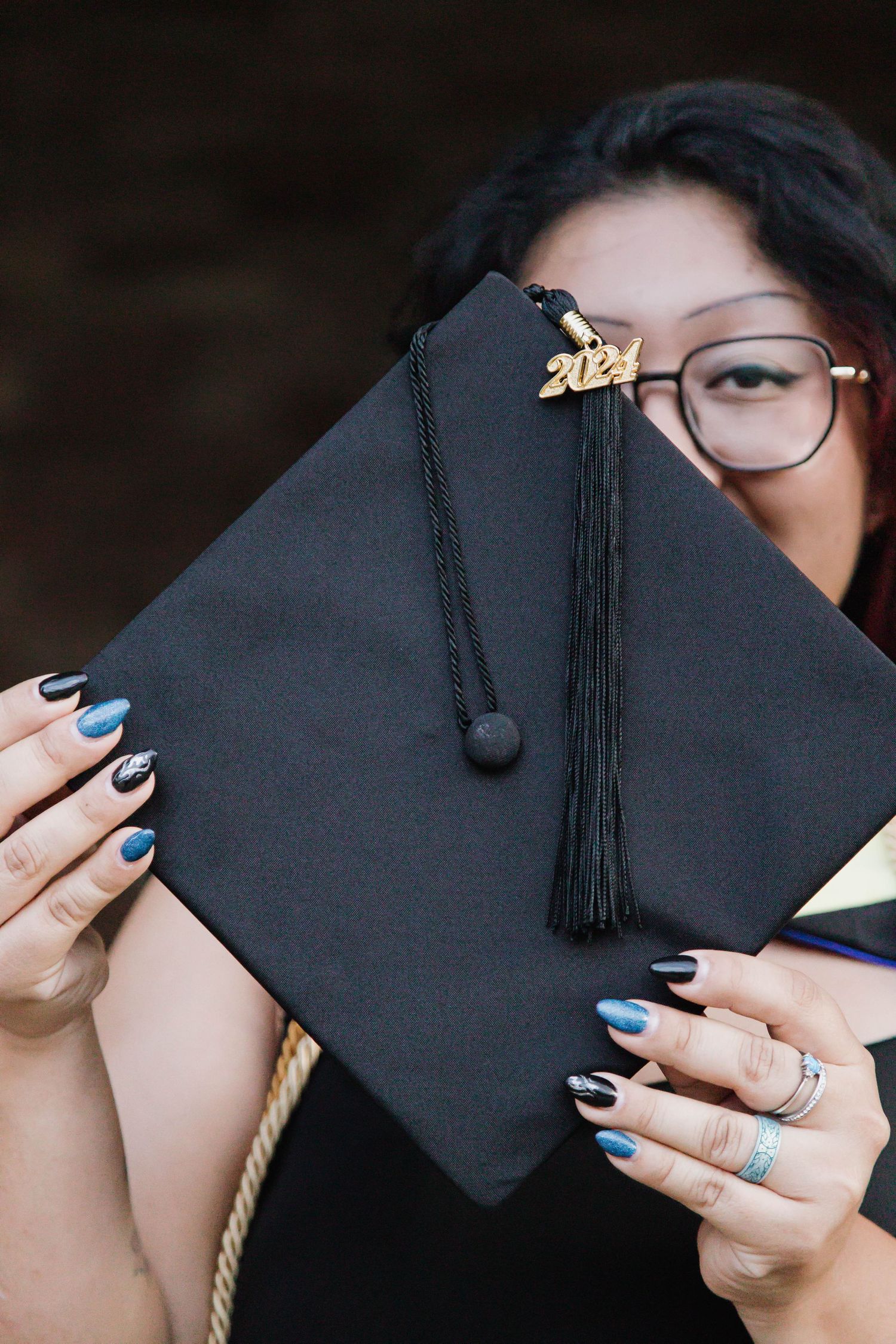 A graduation cap with black tassel held by hands with blue nail polish and glasses visible in the background.