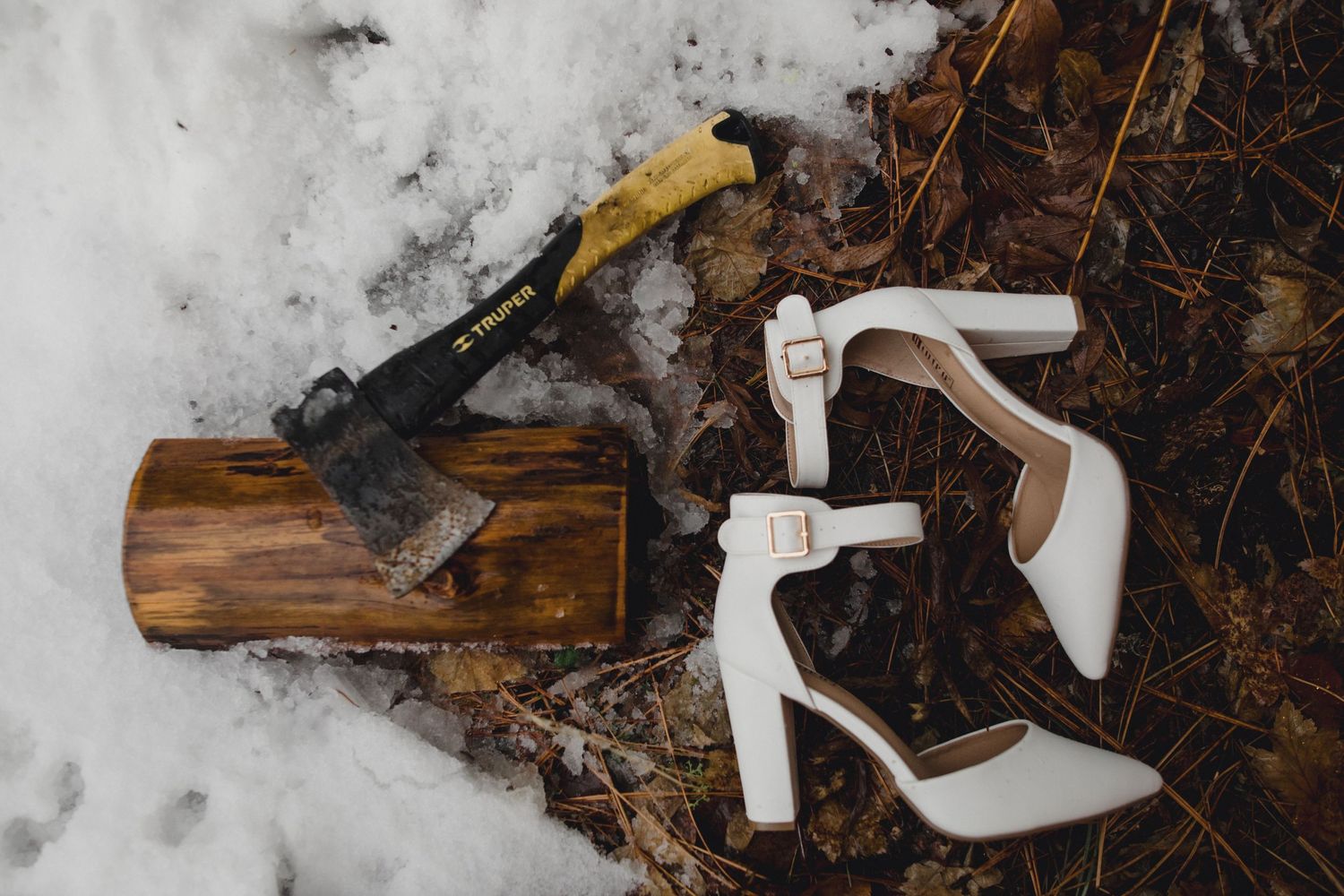 White wedding heels and an axe arranged on a wooden log in snowy winter setting.