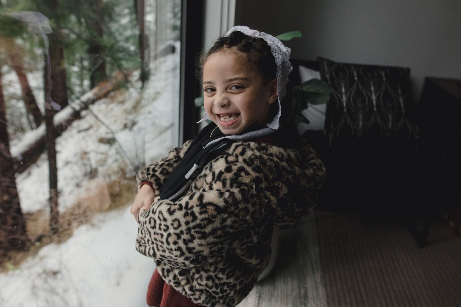 A child in a leopard print winter coat smiles while standing by a snowy window.
