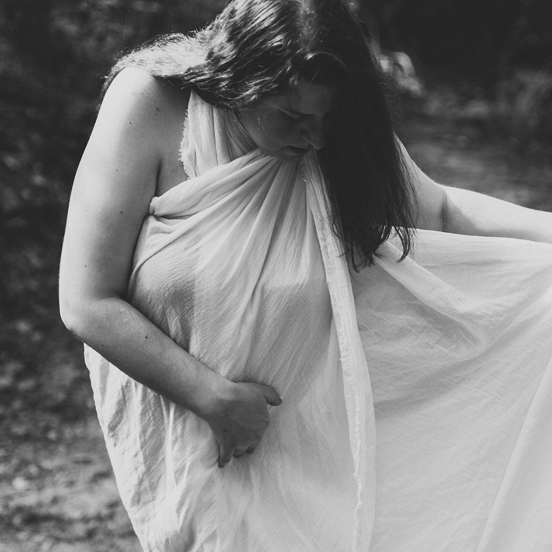Figure draped in flowing white fabric poses artistically outdoors in black and white photography.
