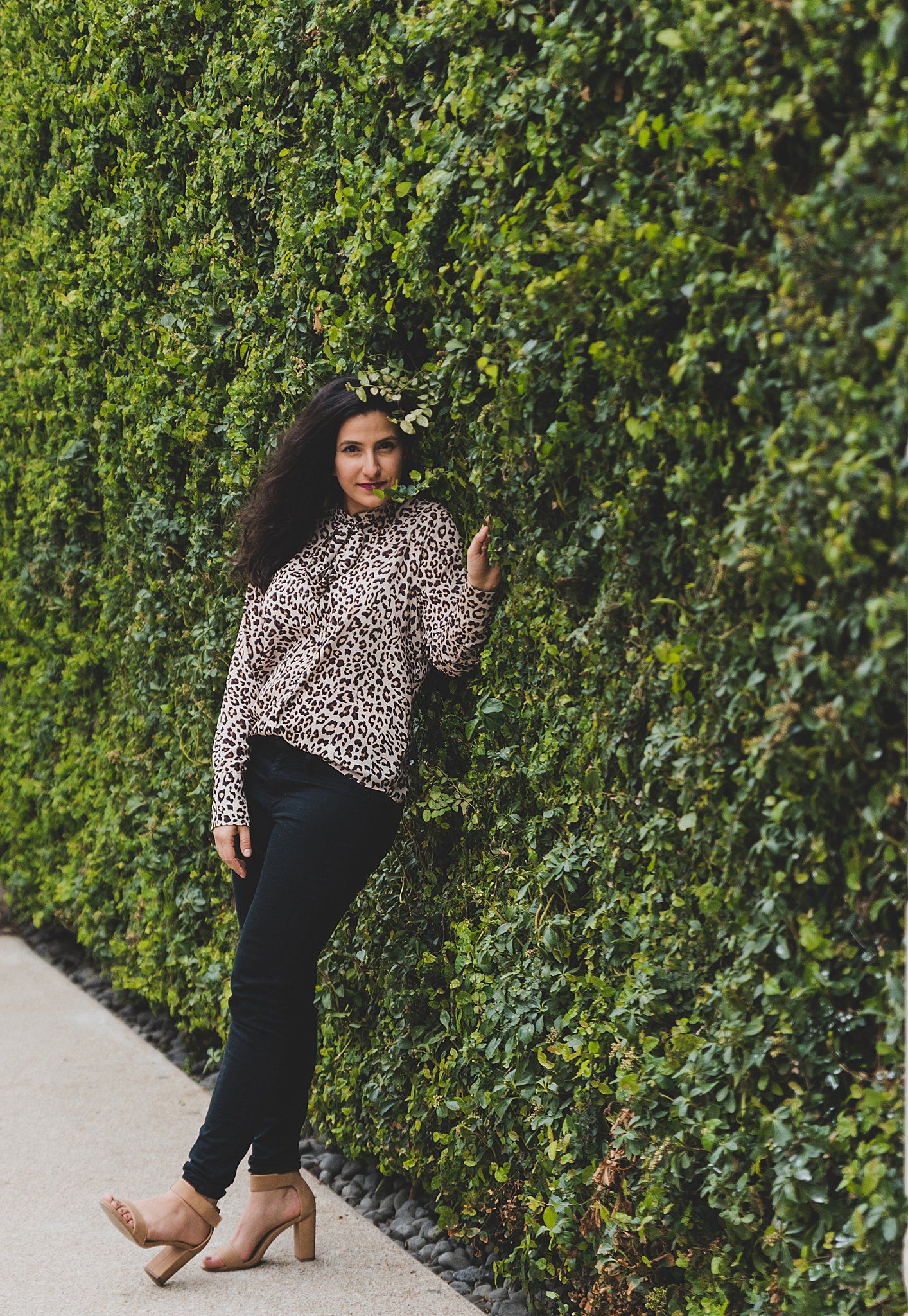 Woman in leopard print top poses against lush green hedge wall.