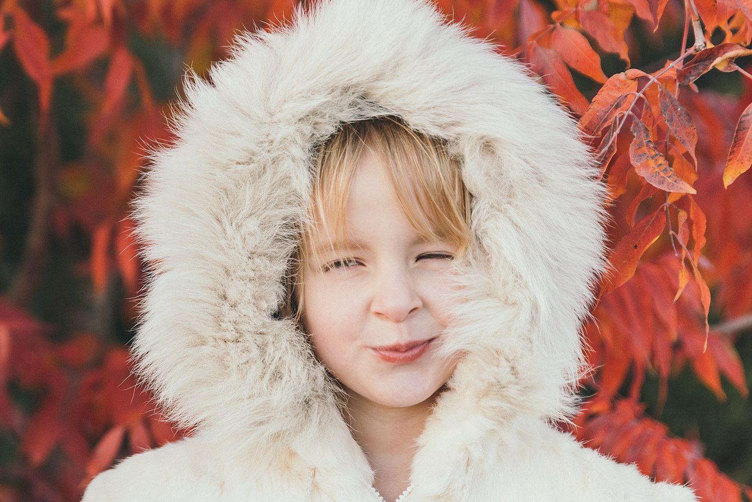 Smiling child in furry white hooded coat against red autumn leaves.