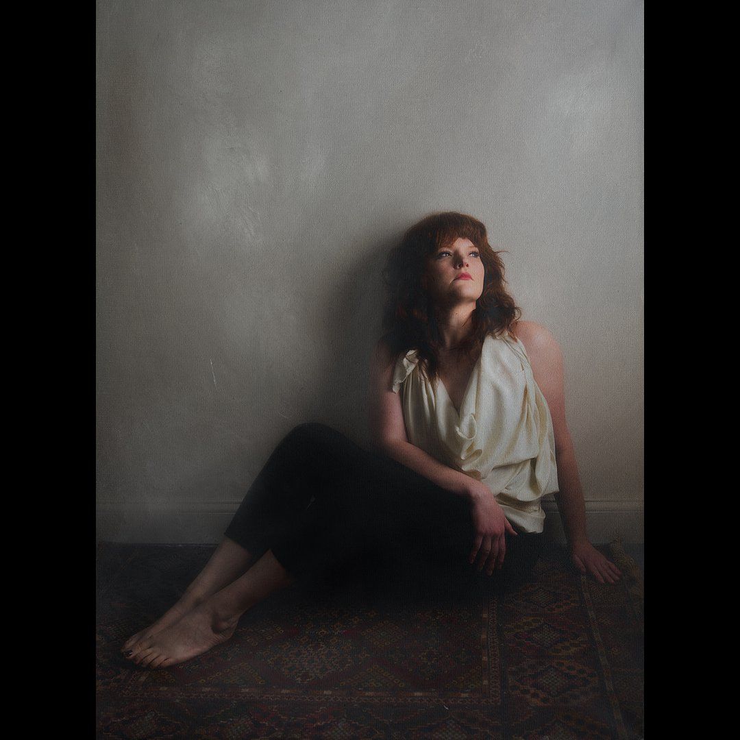 A moody portrait of someone sitting against a wall in a sleeveless top and dark skirt.