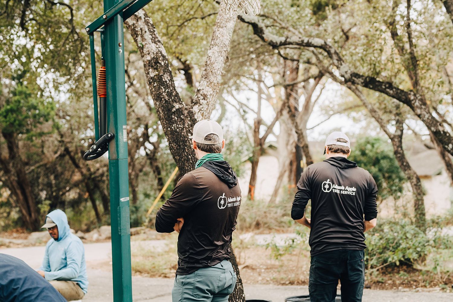 Two people wearing matching black shirts work together on outdoor equipment maintenance in a wooded park area.