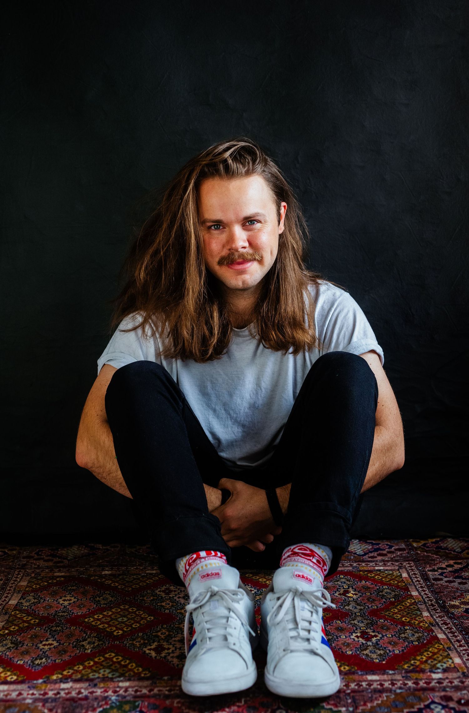 Casual portrait of long-haired person in white t-shirt sitting cross-legged on patterned rug.