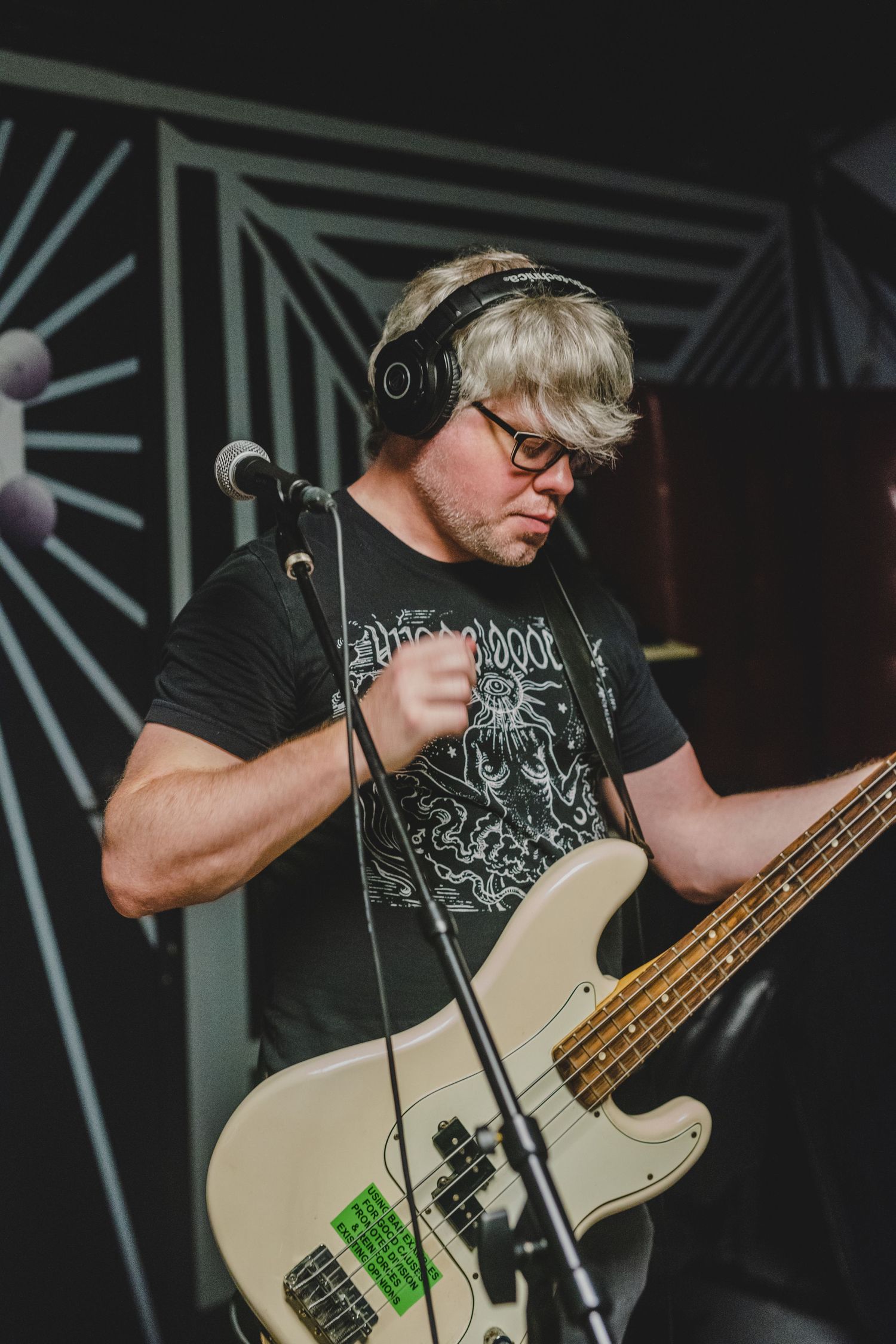 Bassist wearing headphones performs with white guitar against geometric backdrop on stage.