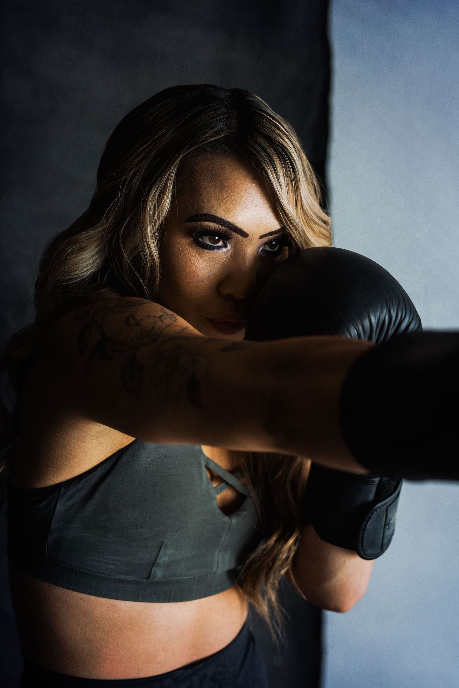 Athletic woman in sports bra and boxing gloves poses dramatically in moody lighting against dark background.