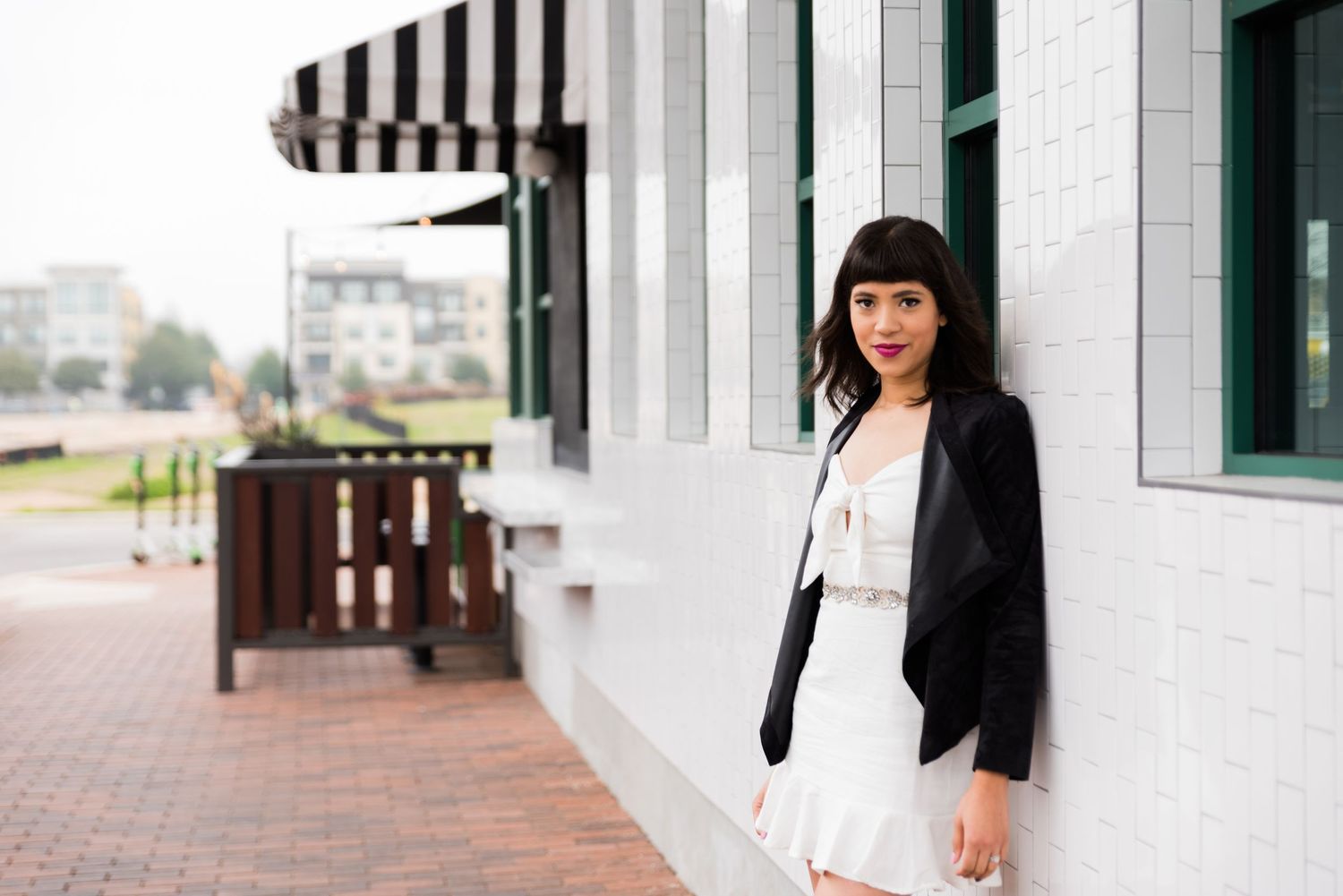 Professional photo of woman in white dress and black jacket standing outside modern building.