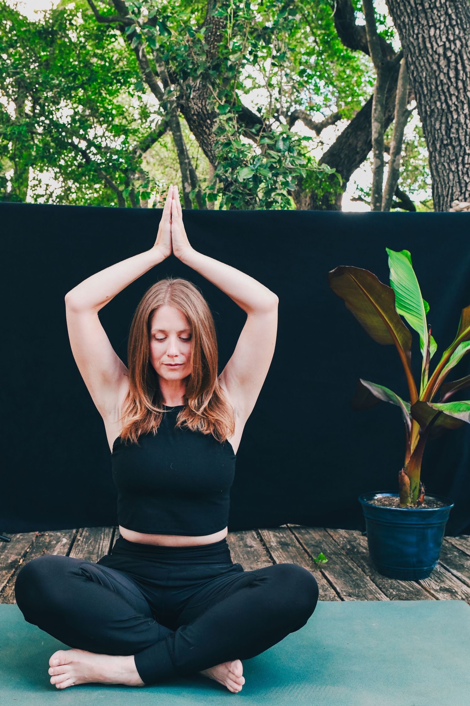 Woman in black activewear practicing yoga meditation pose outdoors with potted plant.