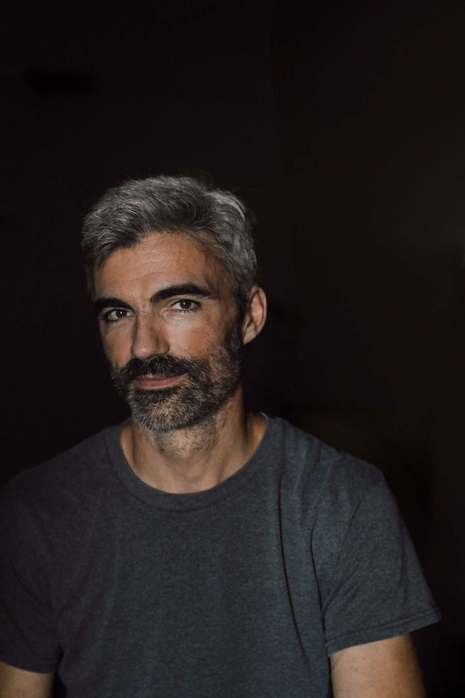 Dramatic low-key portrait of person with salt-and-pepper hair and beard wearing dark t-shirt.