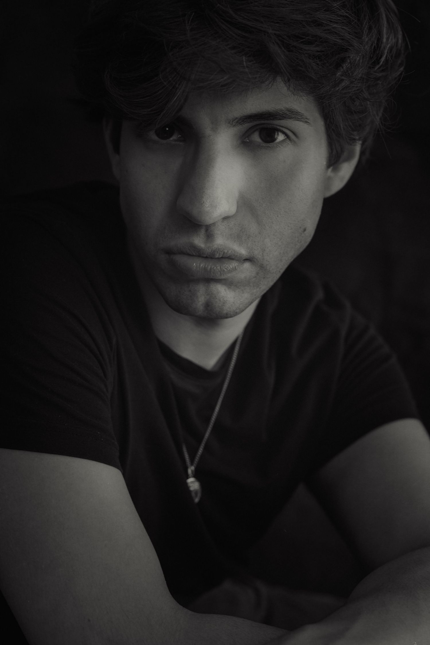 Black and white moody portrait of young person in dark clothing against shadowy background.