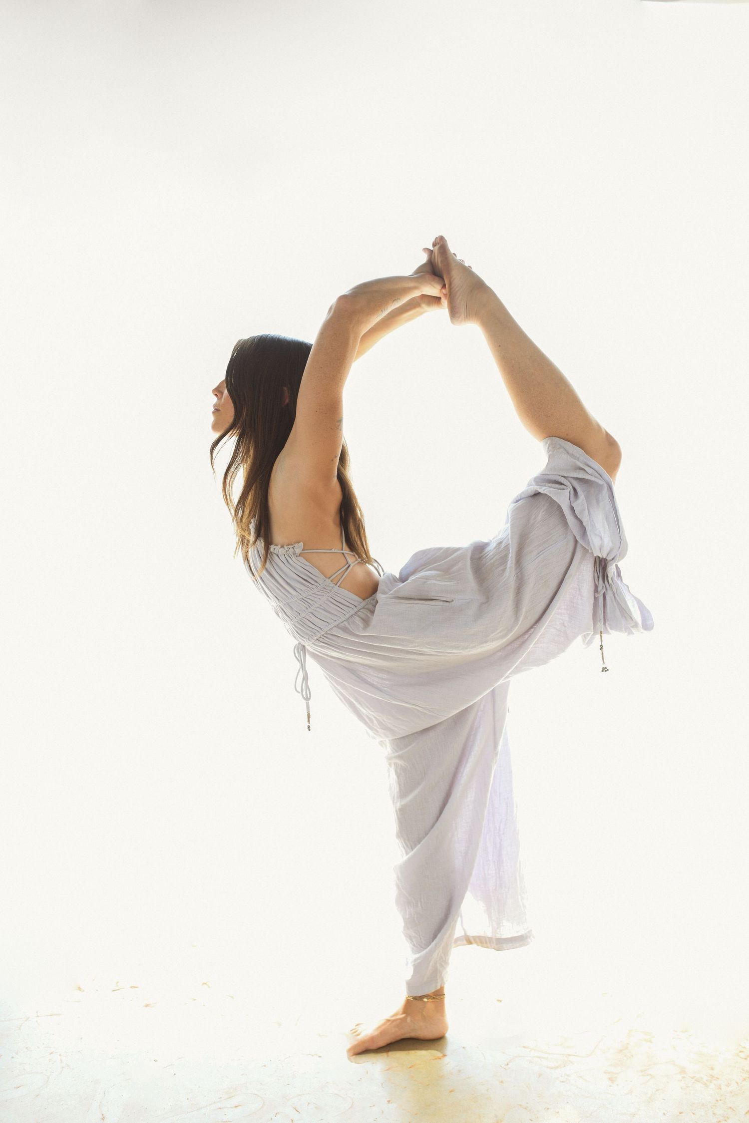 Dancer in flowing gray dress executes perfect standing split pose in bright studio setting.