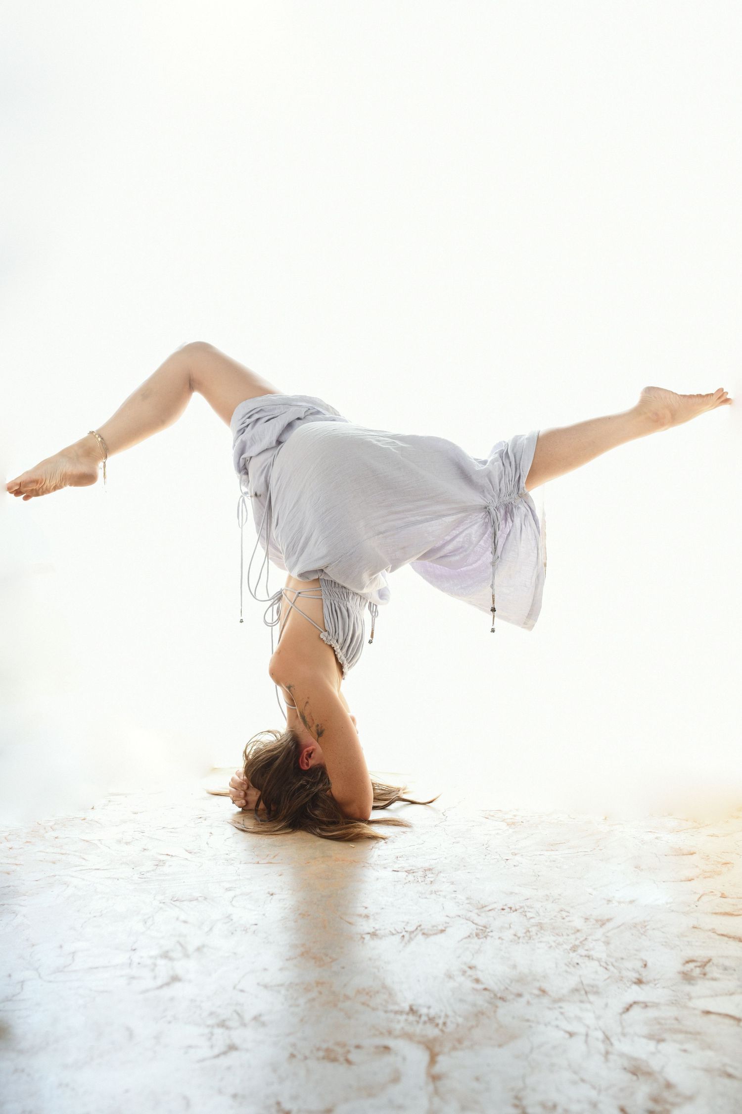Dancer wearing flowing grey dress performs graceful handstand against white background.