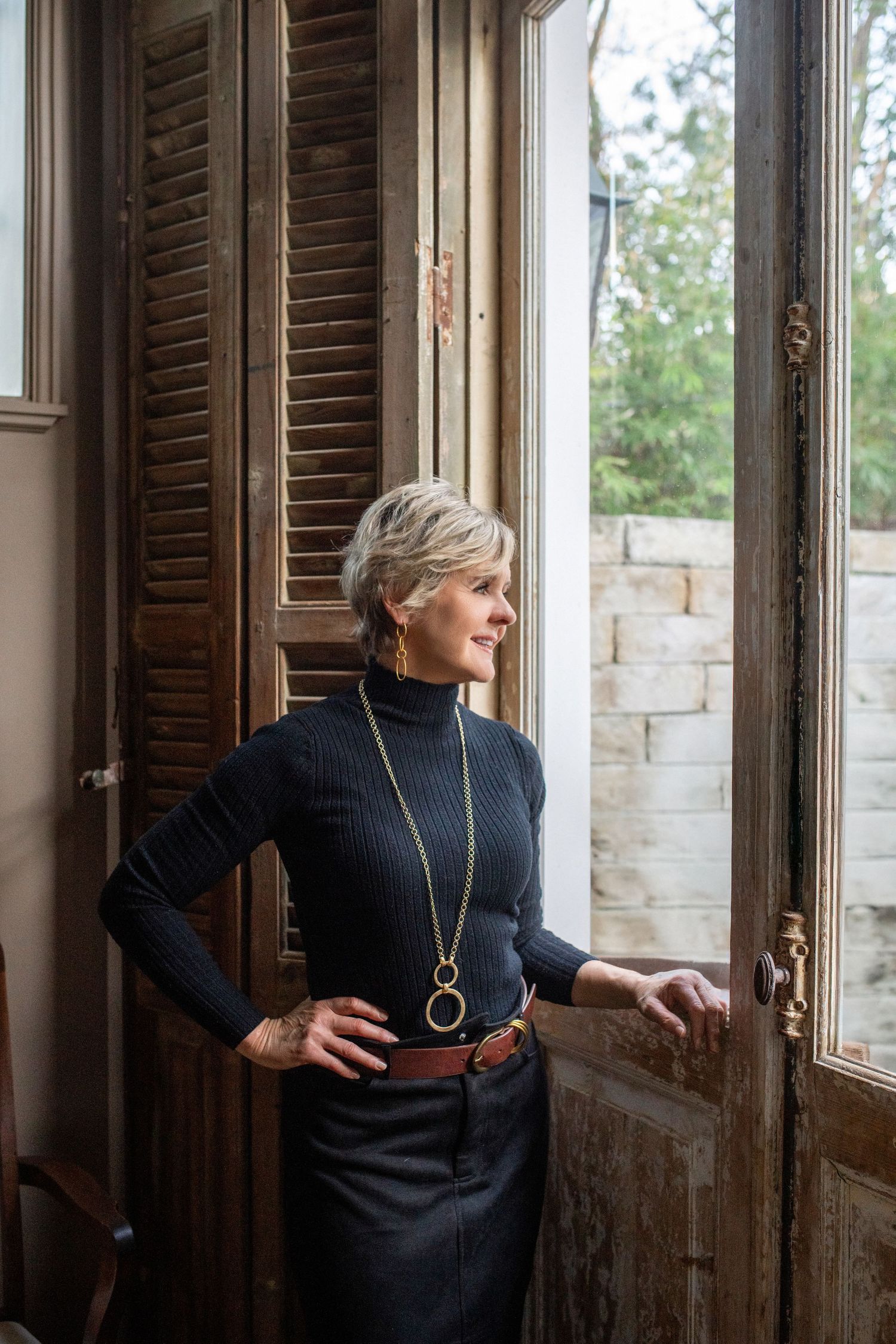 A sophisticated look featuring a black turtleneck sweater and statement necklace by a window with wooden shutters.