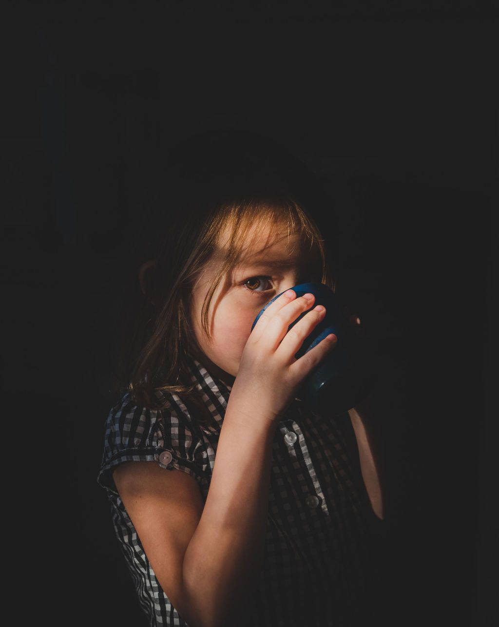 Atmospheric low-key portrait of a child drinking from a cup against dark background.